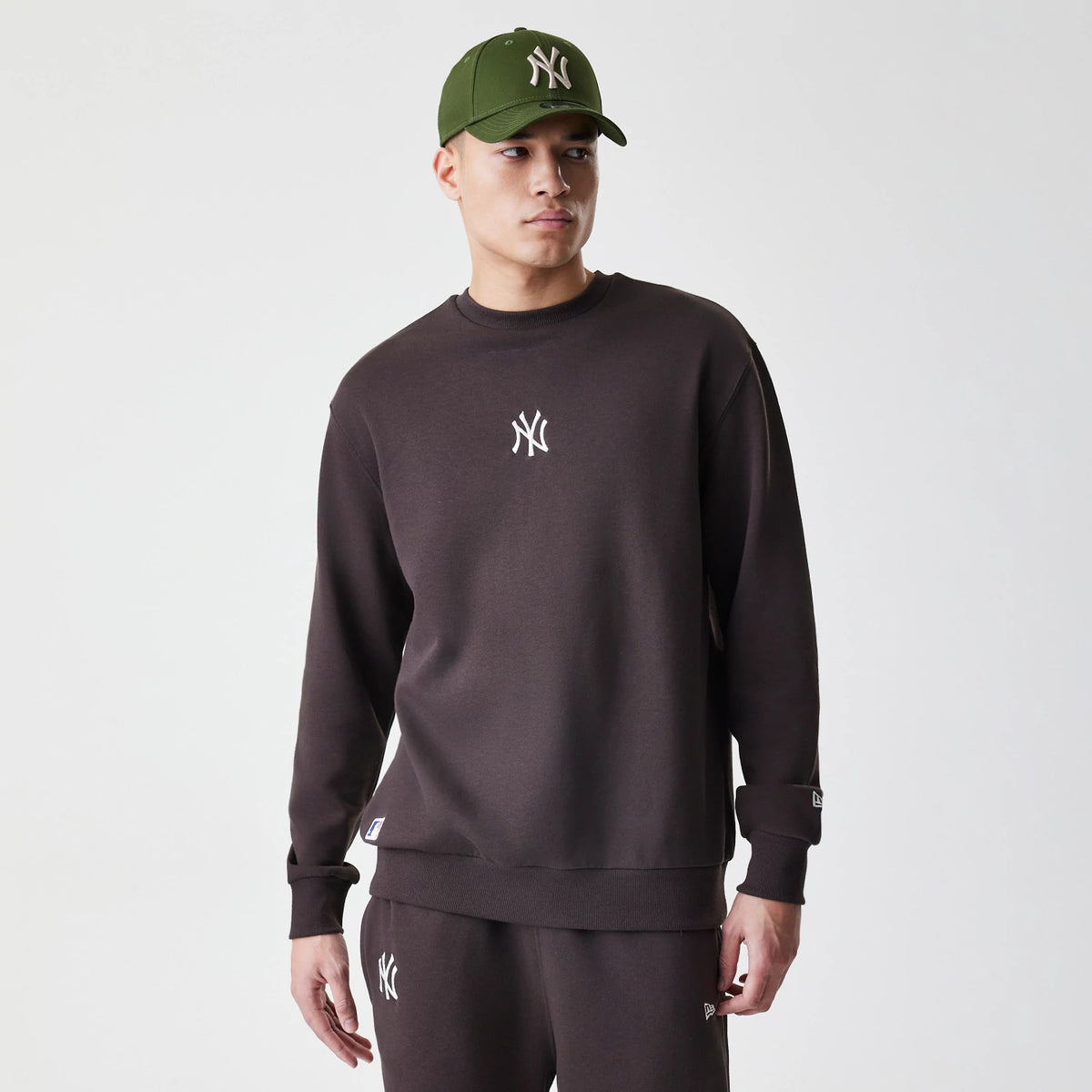 New Era New York Yankees MLB Midi League Essential Dark Brown Oversized Crew Neck Sweater, front view on a model wearing a matching green cap, featuring a white embroidered NY logo on the chest and a New Era flag logo on the sleeve.