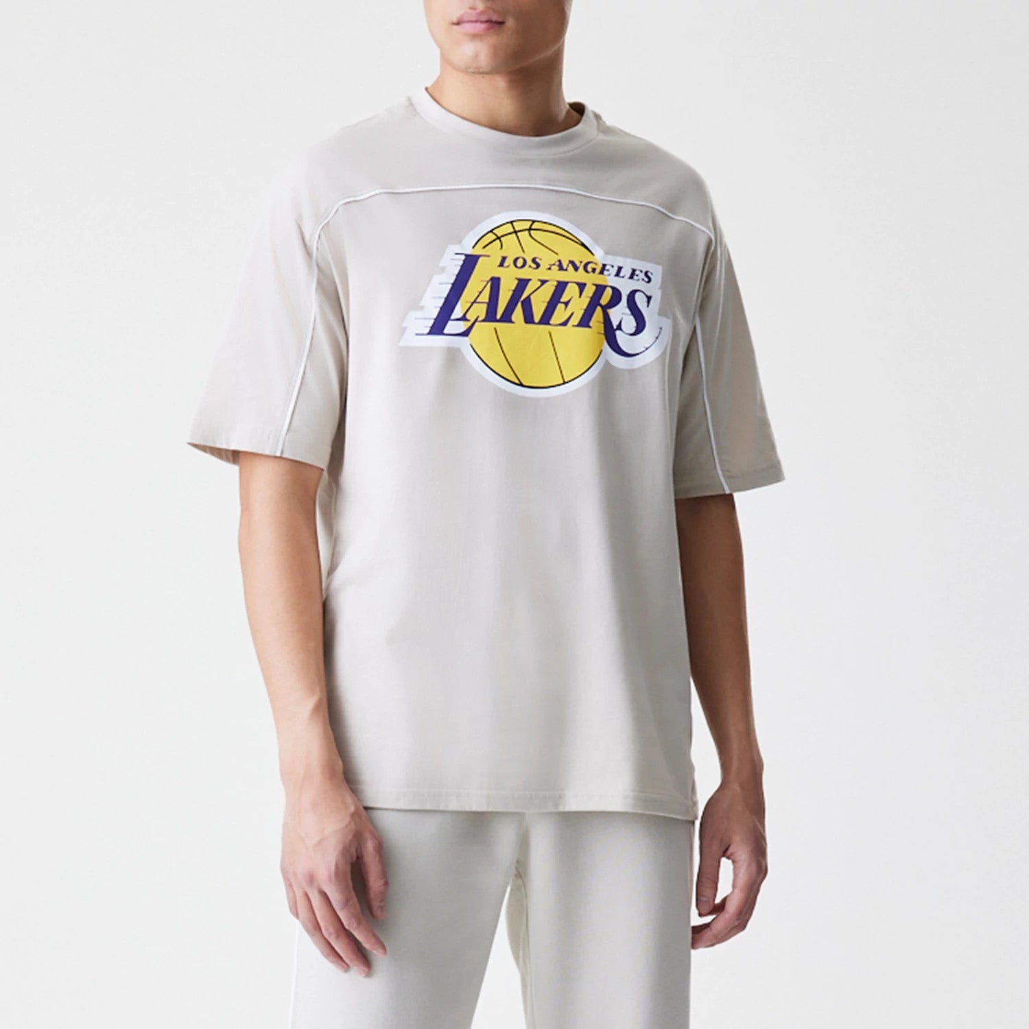 New Era LA Lakers NBA Piping Light Beige Oversized T-Shirt, front view on a model with matching trousers, featuring a large yellow basketball graphic and an embroidered Lakers logo across the chest with white piping.