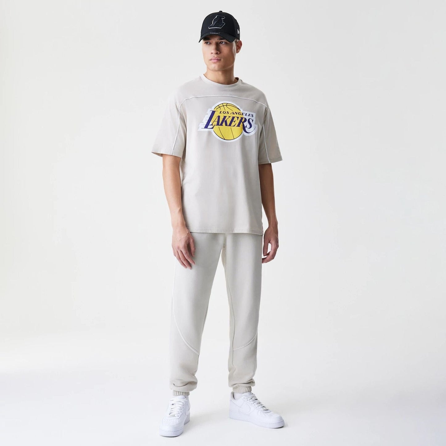 New Era LA Lakers NBA Piping Light Beige Oversized T-Shirt, front view on-model showing the purple and yellow Los Angeles Lakers logo printed across the chest with seam details on the shoulder piping, styled with matching light beige trousers.