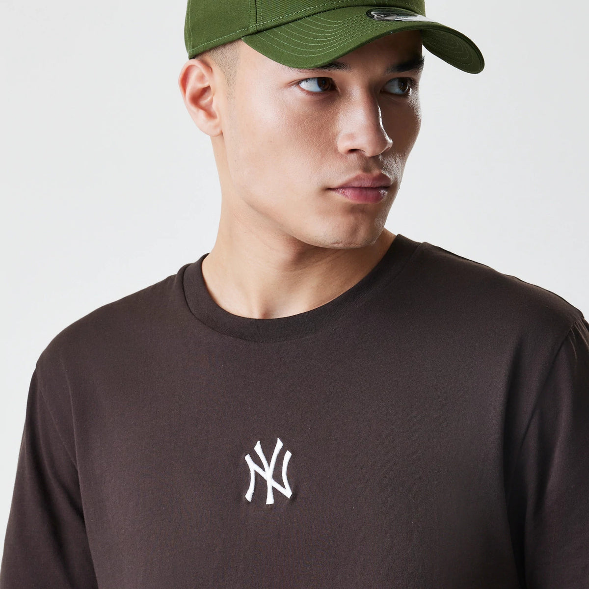 New Era New York Yankees MLB Midi League Essential Dark Brown Oversized T-Shirt, front view showing a white embroidered New York Yankees logo centred on the chest, worn by a model in a green cap.