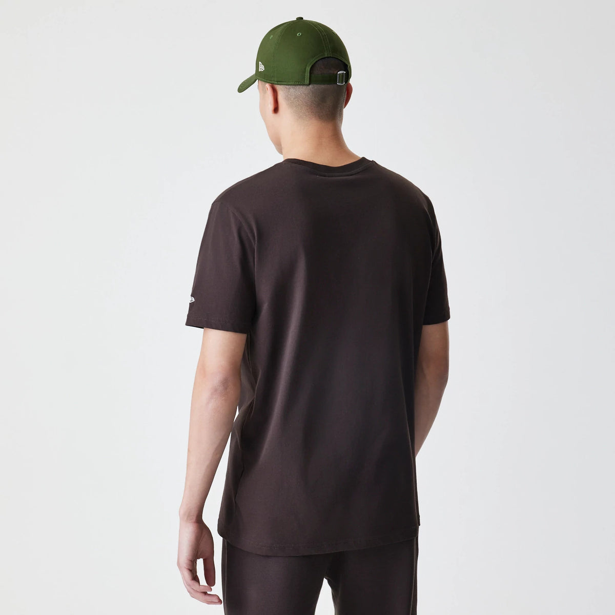 New Era New York Yankees MLB Midi League Essential Dark Brown Oversized T-Shirt, back view on-model showing a solid dark brown back with short sleeves and a small embroidered New Era logo on the left sleeve, paired with a green cap.