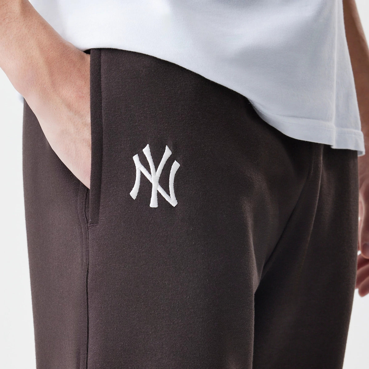 New Era New York Yankees MLB Midi League Essential Dark Brown Joggers, on-model side view with the white embroidered NY logo on the left thigh, showing the elastic waistband and a relaxed fit of the dark brown fabric.