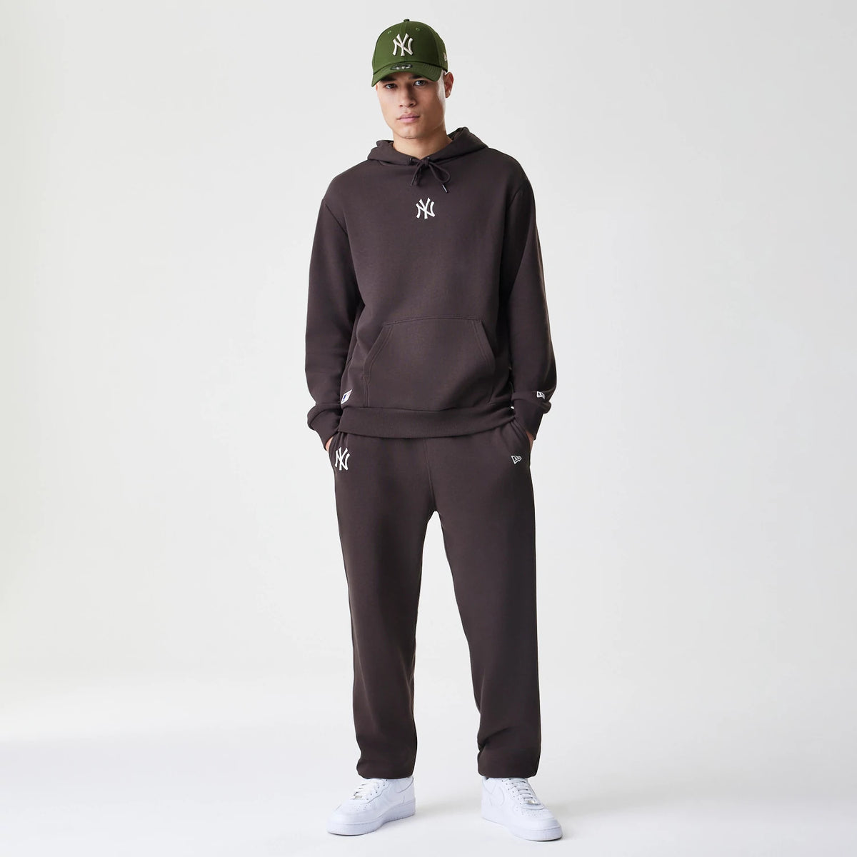 New Era New York Yankees MLB Midi League Essential Dark Brown Joggers, front view on a model paired with a matching hoodie and green cap, showing the white NY logo on the left thigh and New Era flag logo above the left pocket.