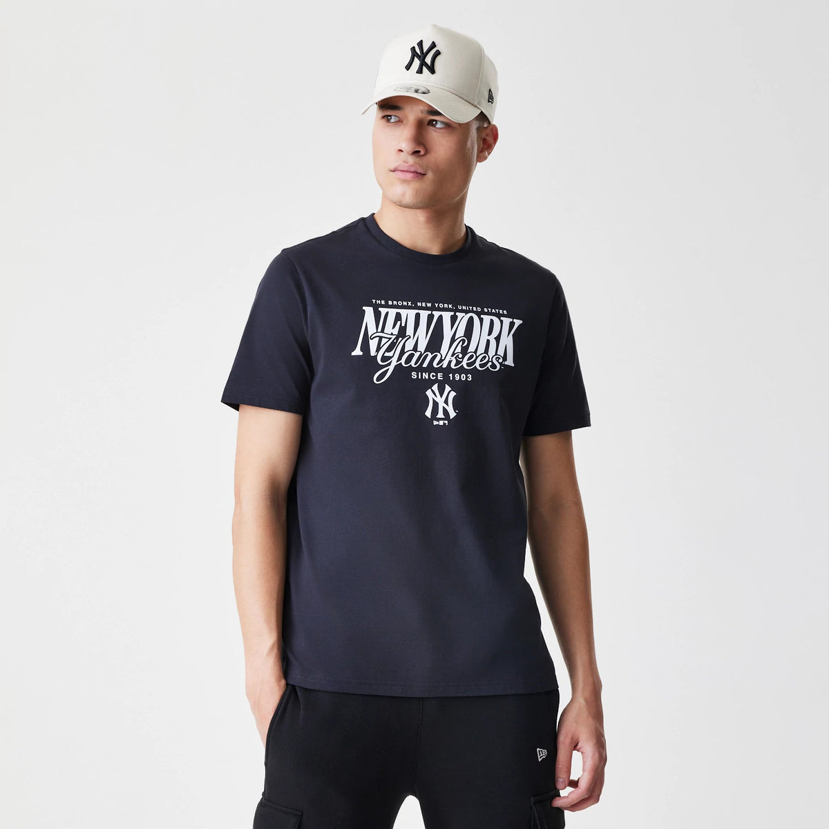 New Era New York Yankees MLB Script Graphic Navy T-Shirt, front view on-model displaying white New York Yankees script across the chest with a small MLB logo beneath, paired with a cream cap featuring an embroidered Yankees logo.