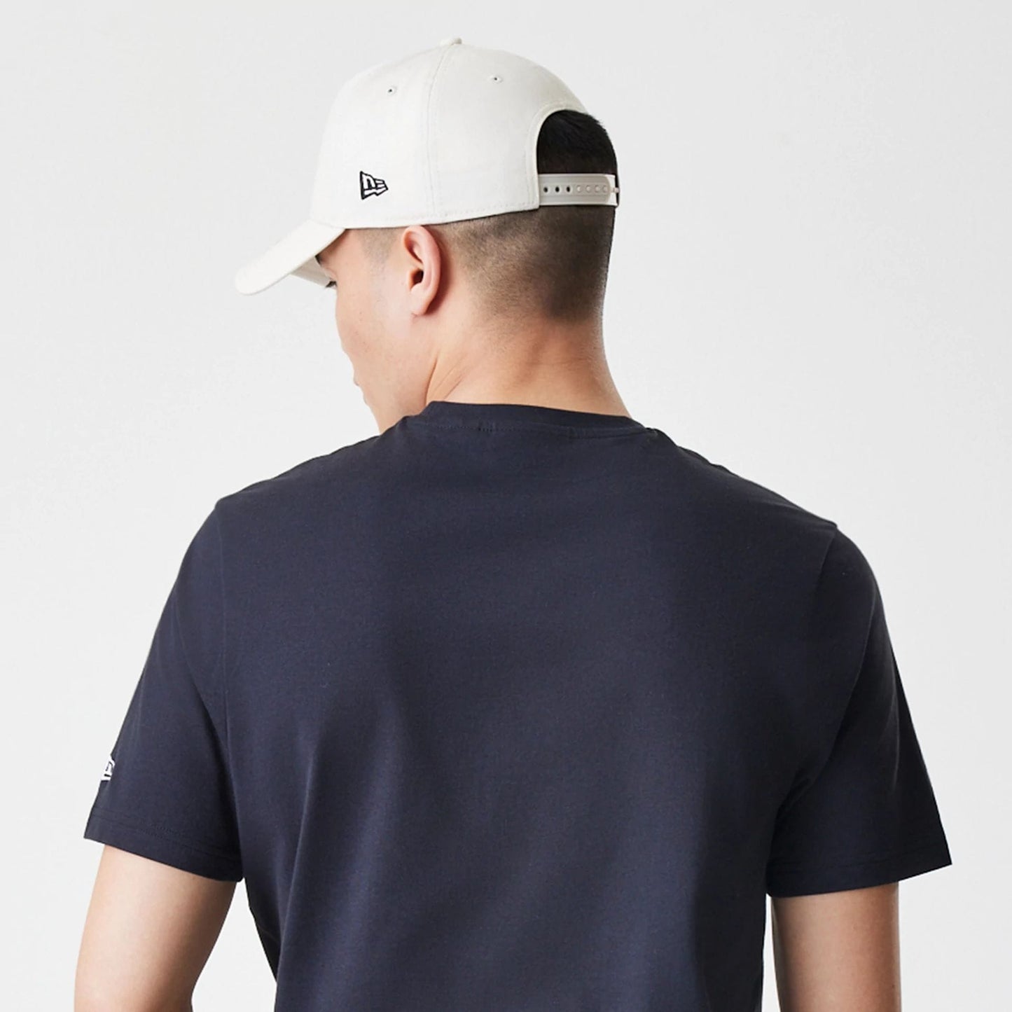 New Era New York Yankees MLB Script Graphic Navy T-Shirt, back view on-model showing the plain navy back of the shirt with short sleeves, an embroidered New Era logo on the left sleeve, paired with a white cap featuring an embroidered side logo.