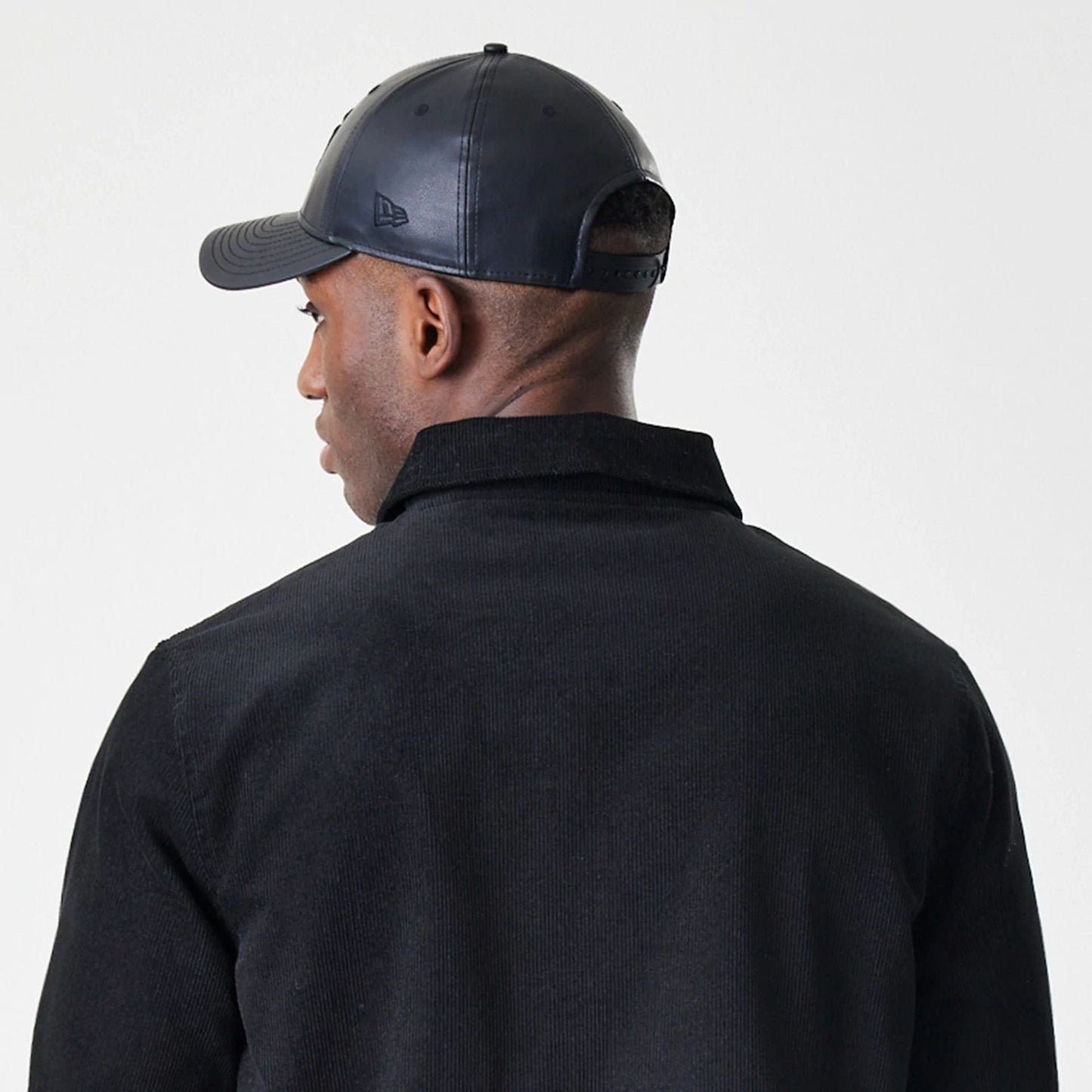 New Era Cord Black Coaches Jacket, back view on a model wearing a matching black cap, showing the simple black cord design with a structured collar and clean lines running along the shoulder seams.