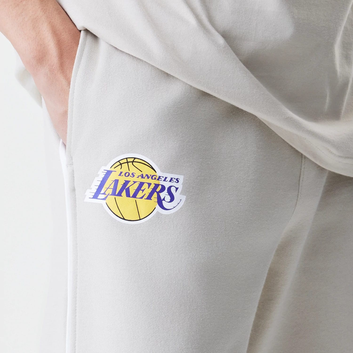 New Era LA Lakers NBA Piping Cream Joggers, close-up showing the Los Angeles Lakers purple and yellow basketball logo embroidered near the right pocket on a cream fabric, highlighting the soft texture and relaxed fit.