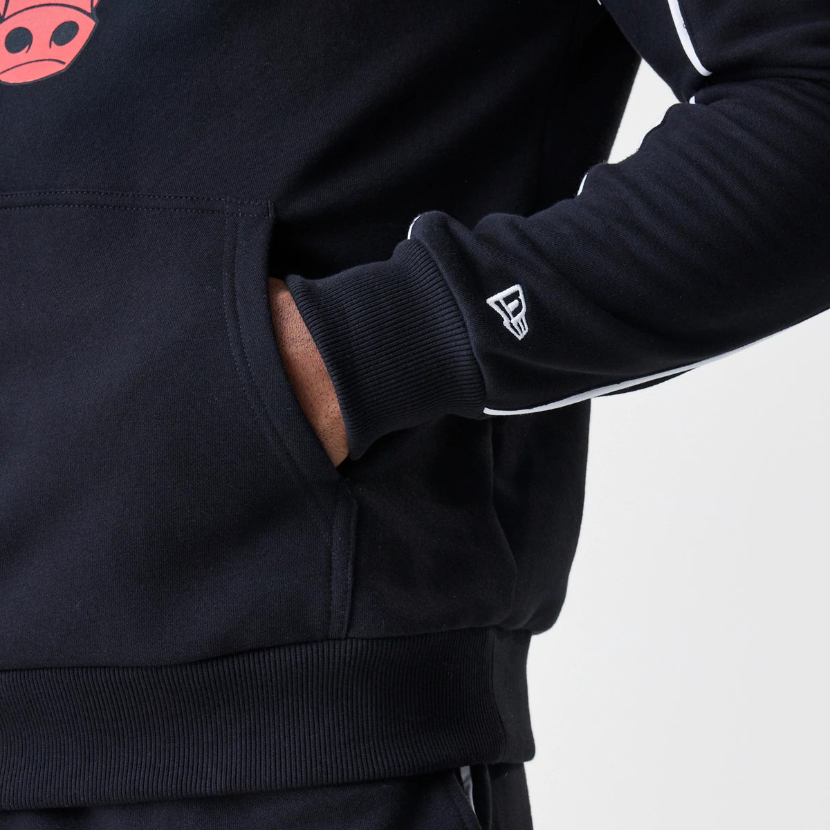 New Era Chicago Bulls NBA Piping Stripe Black Oversized Pullover Hoodie, side view showing the embroidered New Era logo on the sleeve cuff, white piping along the arm, and a glimpse of the red Bulls graphic on the chest.