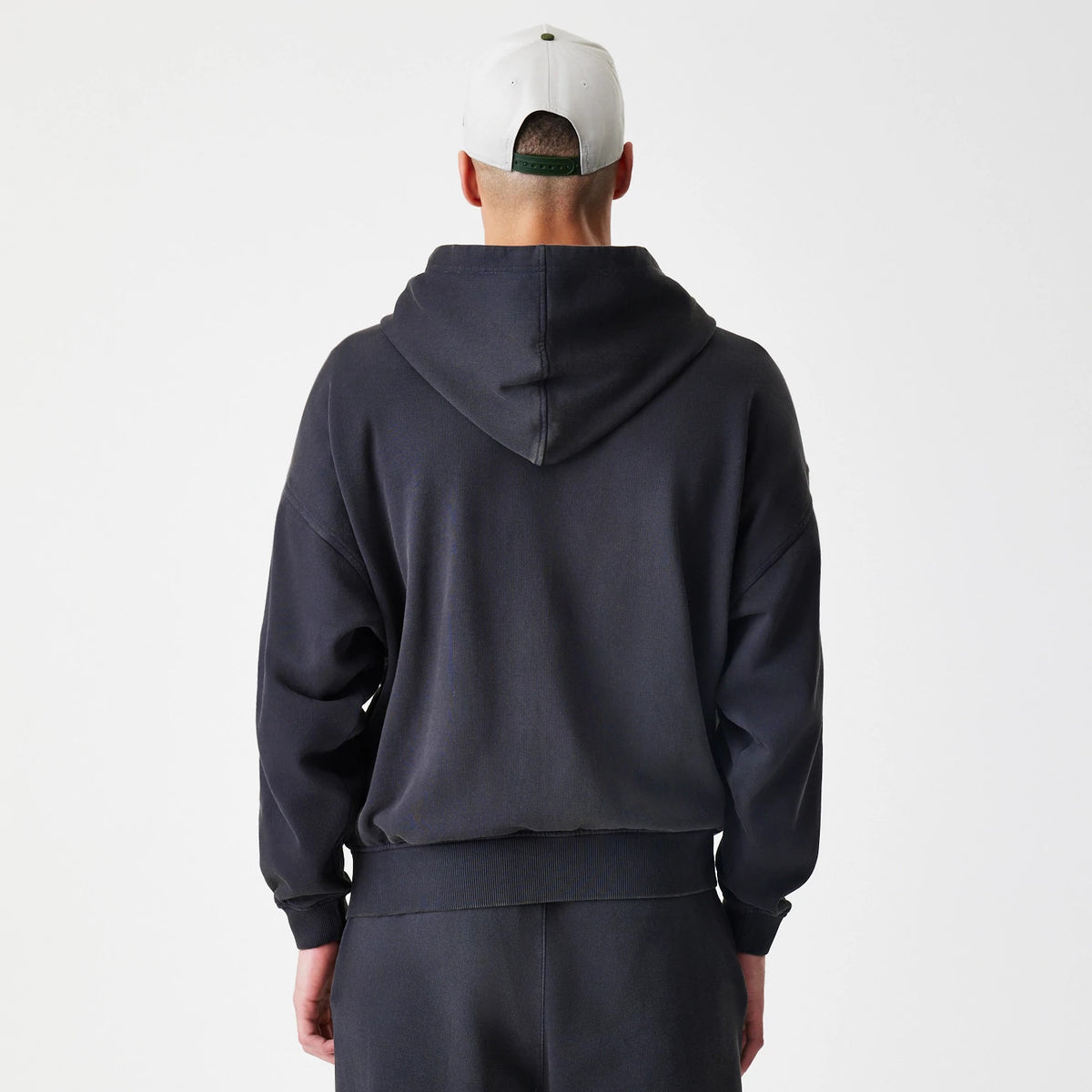 New Era Washed Black Oversized Full Zip Hoodie, back view showing a relaxed fit with a large hood and ribbed cuffs and hem, styled with plain matching trousers and a contrasting cap.