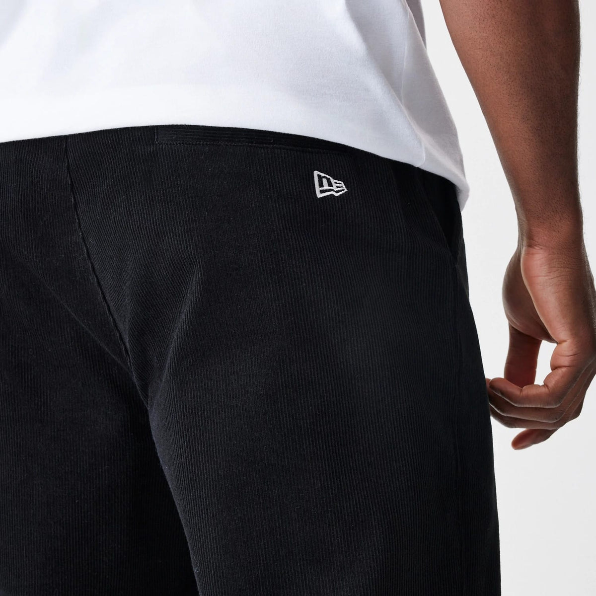 New Era Cord Black Straight Leg Chinos, rear view on a model showing the fine corduroy texture and the white embroidered New Era flag logo on the right side below the waist, paired with a white top.