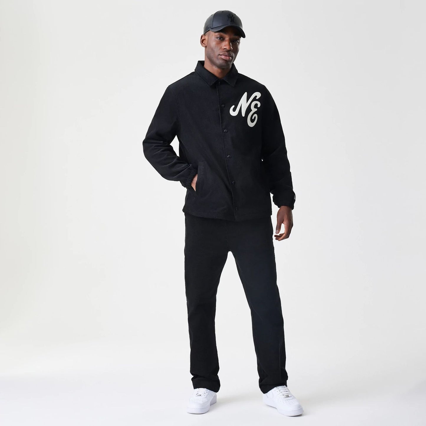New Era Cord Black Straight Leg Chinos, on-model view showing the full fit with a relaxed silhouette, paired with a matching black jacket featuring large white New Era lettering on the chest.