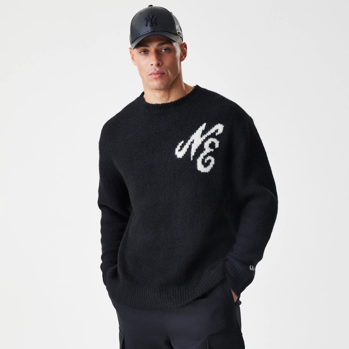 New Era Knit Black Knitted Sweater, front view on a model wearing a black cap, featuring large white scripted NE lettering on the left chest and ribbed cuffs, with the New Era flag logo on the left sleeve.