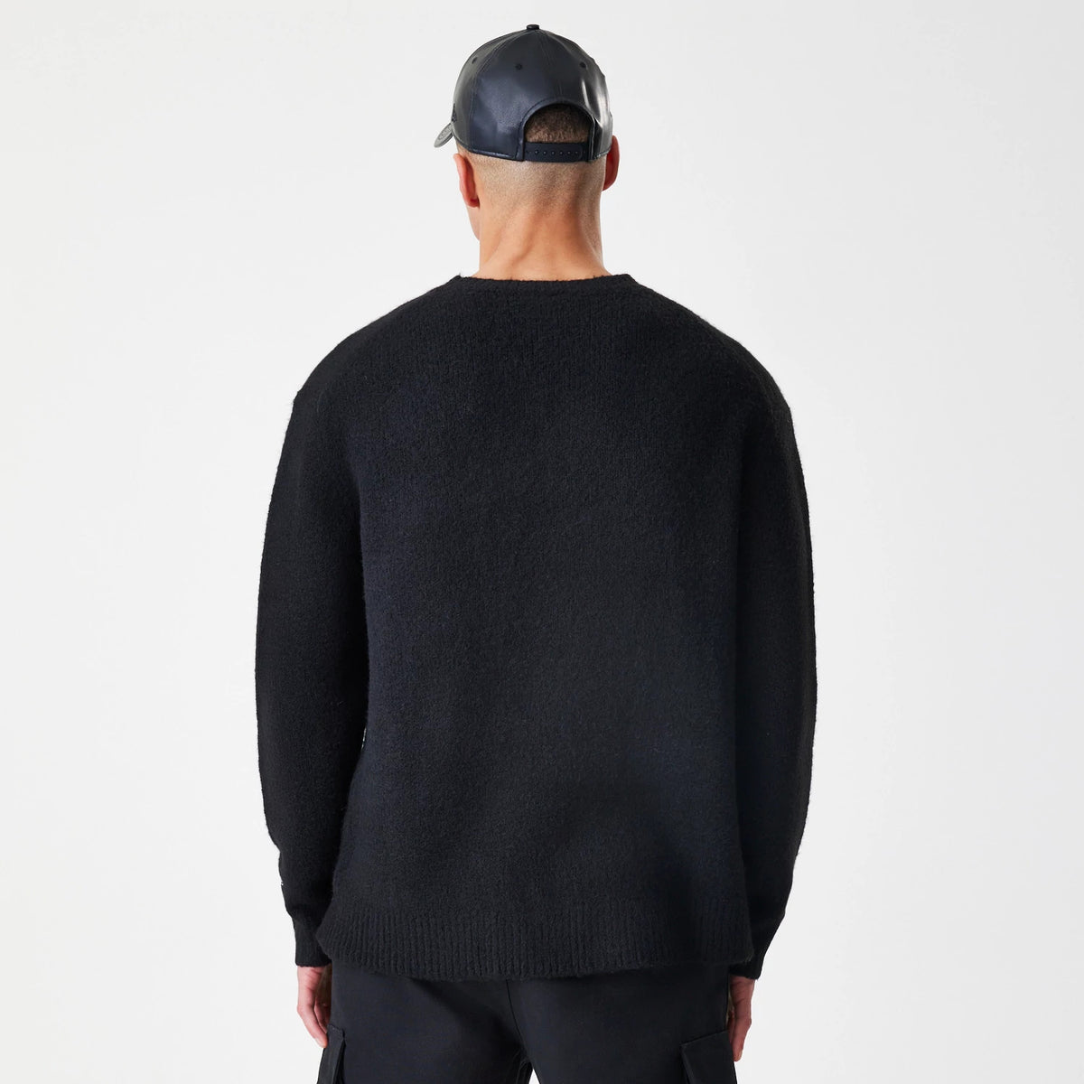 New Era Knit Black Knitted Sweater, back view on a model wearing a black cap, showing the sweater's simple design with a clean neckline and long sleeves, highlighting the ribbed hem and relaxed fit.
