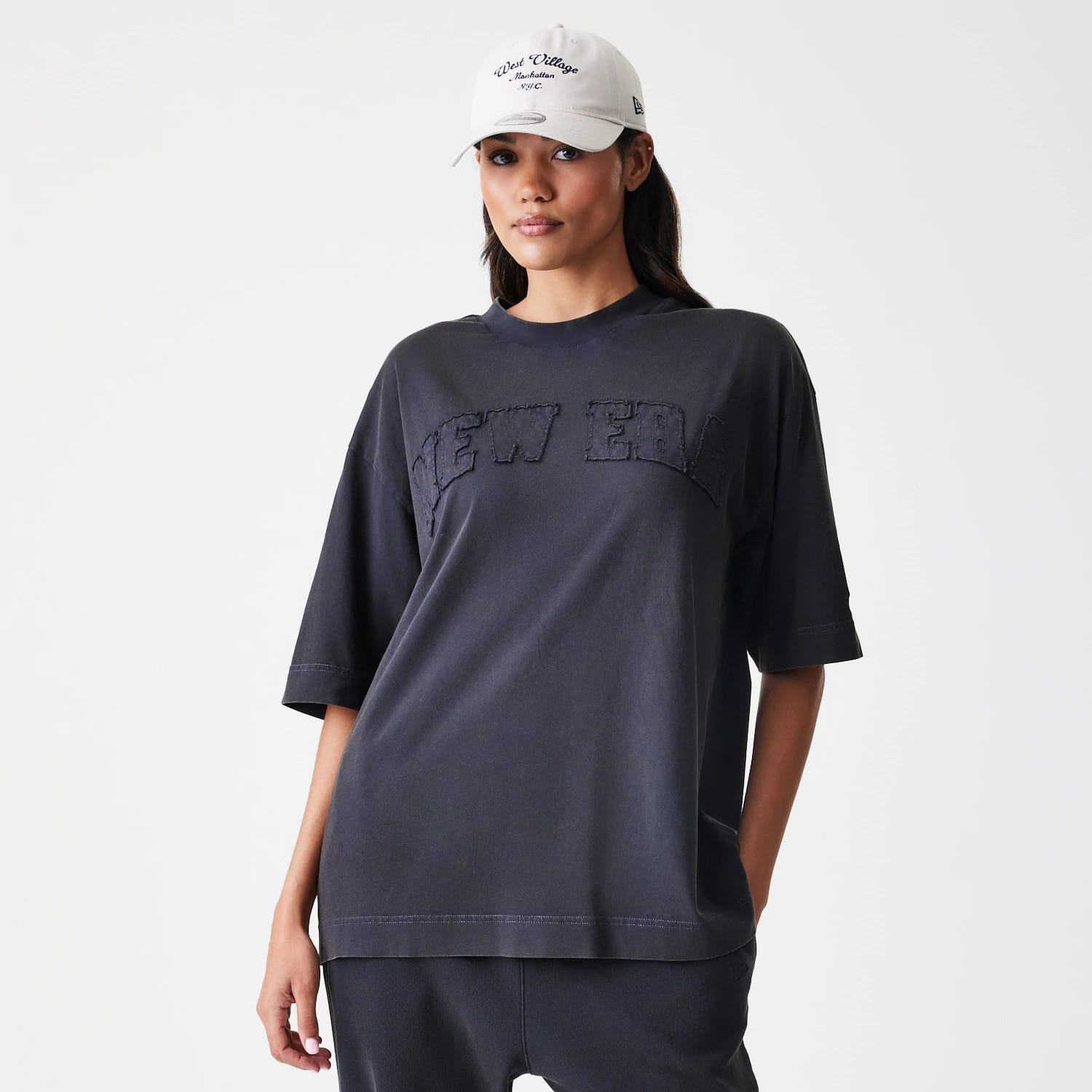 New Era Washed Black Oversized T-Shirt, front view on a model wearing a light-coloured cap, featuring large raised New Era lettering across the chest on a loose-fitting washed black fabric.