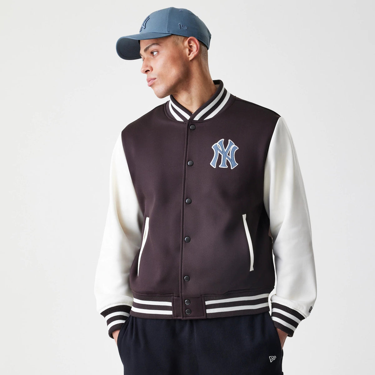 New Era New York Yankees MLB Dark Brown Varsity Jacket, front view on-model showing the large blue and white embroidered NY logo on the chest, with contrasting white sleeves and black and white striped ribbed collar, cuffs, and hem.