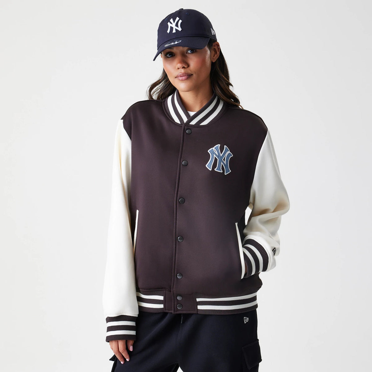 New Era New York Yankees MLB Dark Brown Varsity Jacket, front view on a model wearing a matching cap, displaying a blue and white embroidered New York Yankees logo on the chest and striped ribbed cuffs and collar.