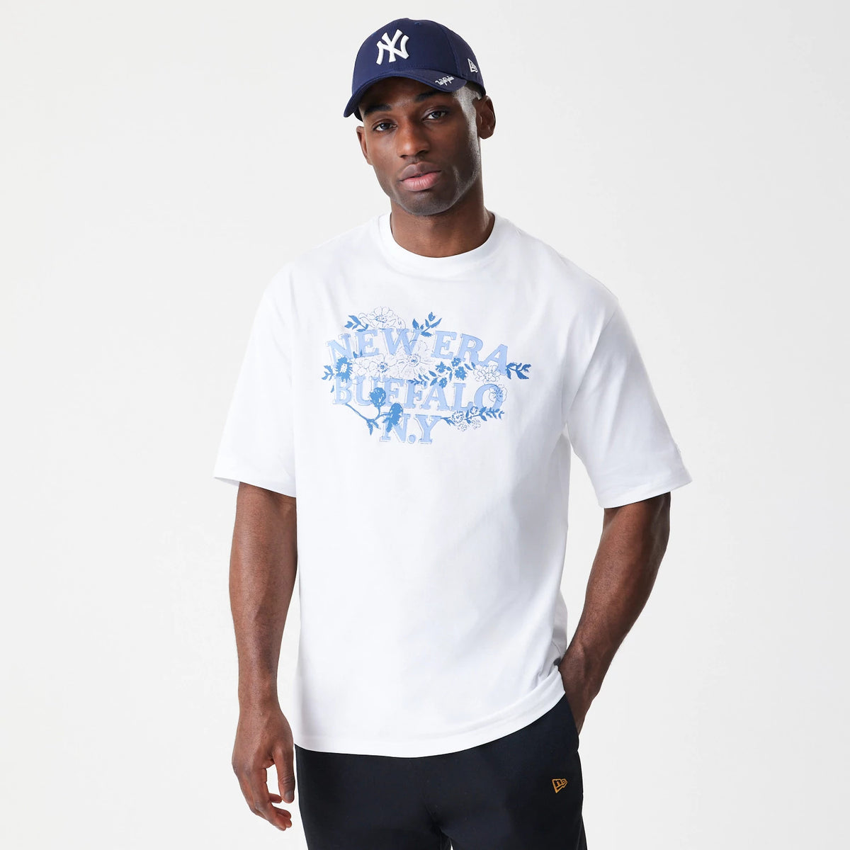 New Era Floral White Oversized T-Shirt, front view on-model showing the blue floral graphic with New Era Buffalo N.Y. lettering across the chest, paired with black trousers and a dark New Era cap.
