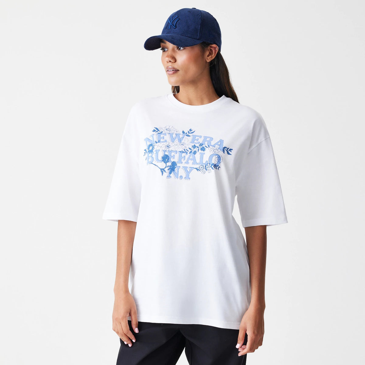 New Era Floral White Oversized T-Shirt, front view on-model showing large blue floral graphic with 'New Era Buffalo N.Y' text across the chest, styled with a dark blue cap and contrasting black trousers.