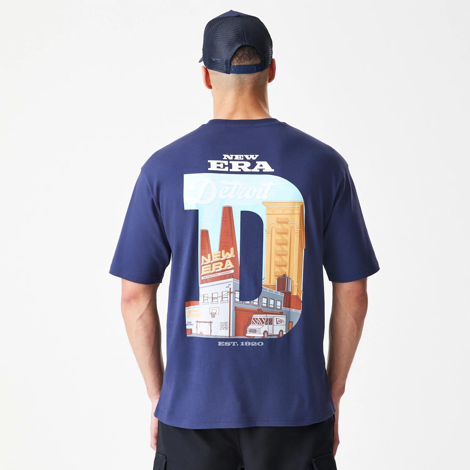 New Era Location Navy Oversized T-Shirt, back view on a model featuring a large graphic of the letter D with Detroit cityscape elements, New Era lettering above and EST. 1920 below, styled with a mesh cap.