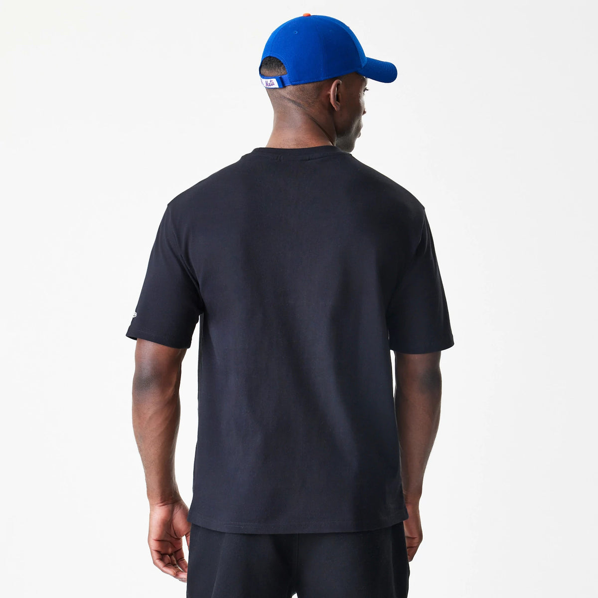 New Era New York Mets MLB Script Black Oversized T-Shirt, back view on a model with a blue cap, showing the plain black back with short sleeves and a small embroidered New Era logo on the left sleeve.