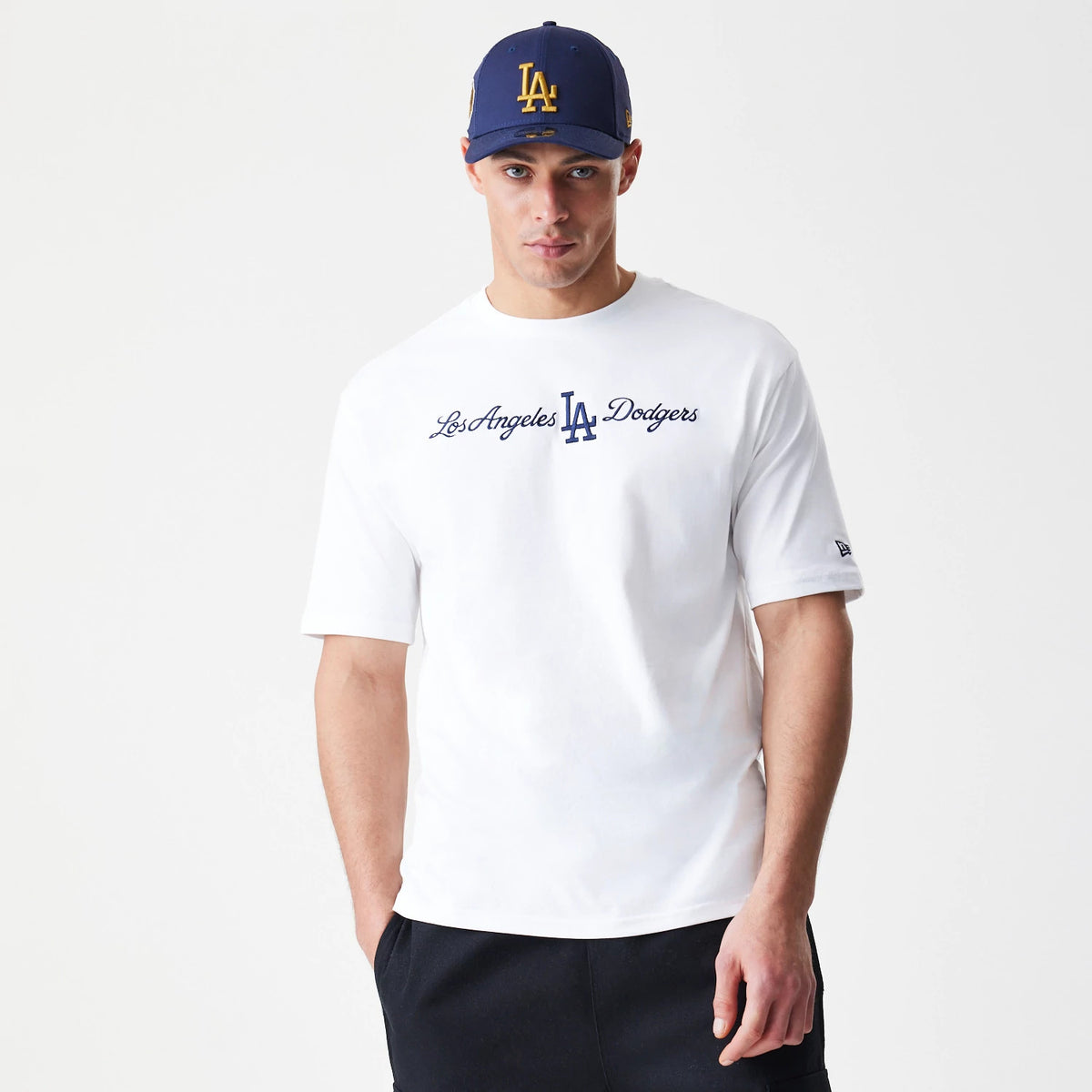 New Era LA Dodgers MLB Script White Oversized T-Shirt, front view on a model with a navy cap, displaying blue Los Angeles Dodgers script across the chest and a small embroidered New Era logo on the left sleeve.