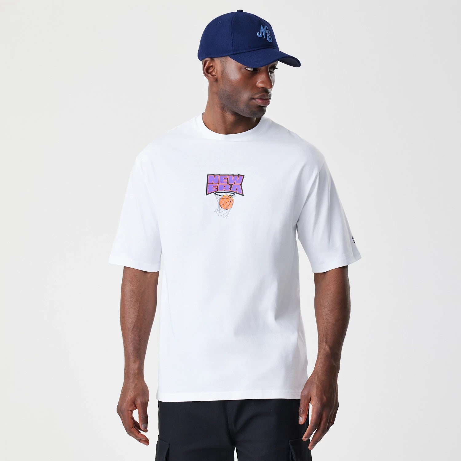 New Era Location White Oversized T-Shirt, front view on-model showing a colourful basketball graphic with the New Era logo printed on the chest, styled with a dark blue cap featuring an embroidered NE on the front.
