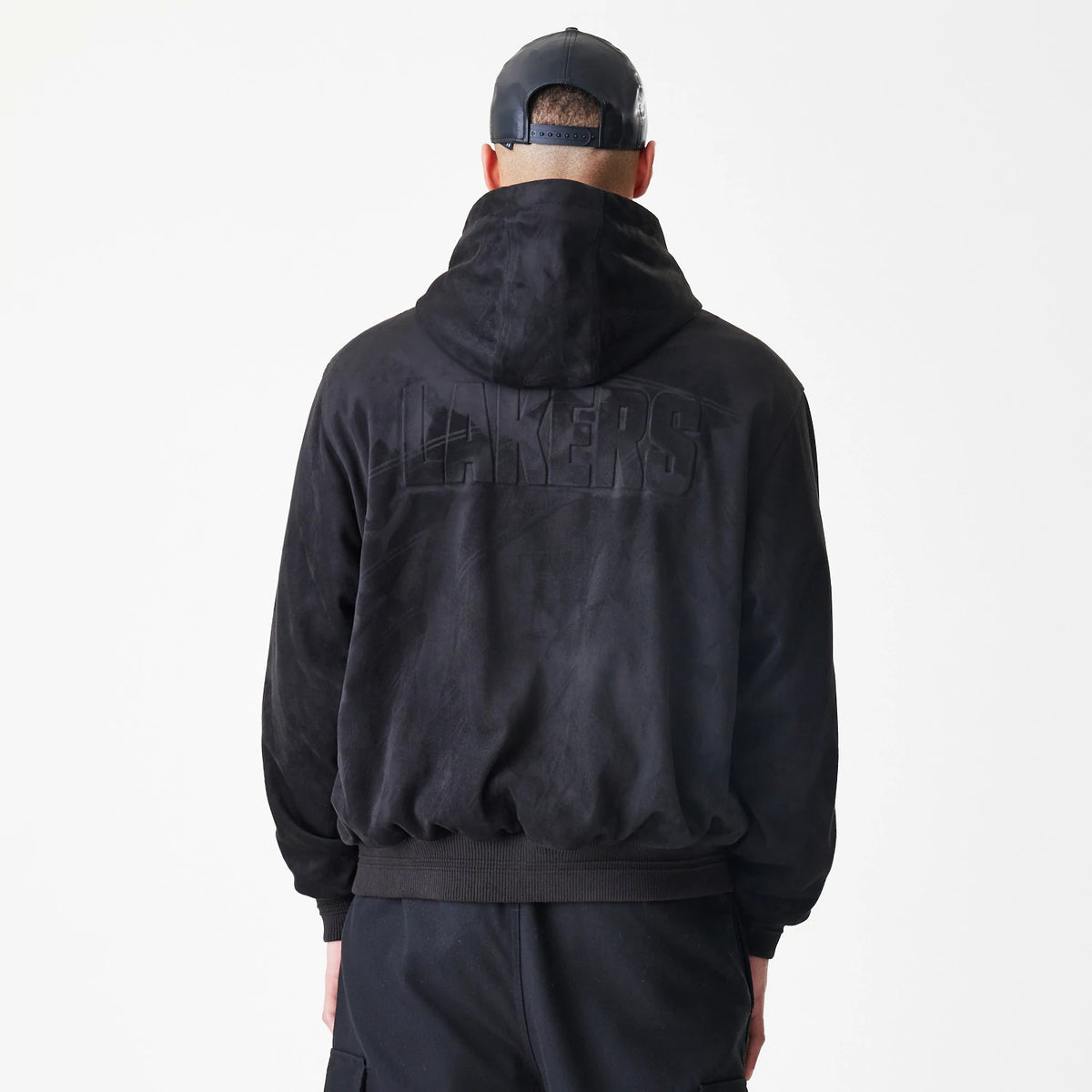 New Era LA Lakers NBA Suede Black Hooded Jacket, back view on a model wearing a matching cap, showing large raised Lakers lettering across the upper back beneath the hood of the plush black jacket.