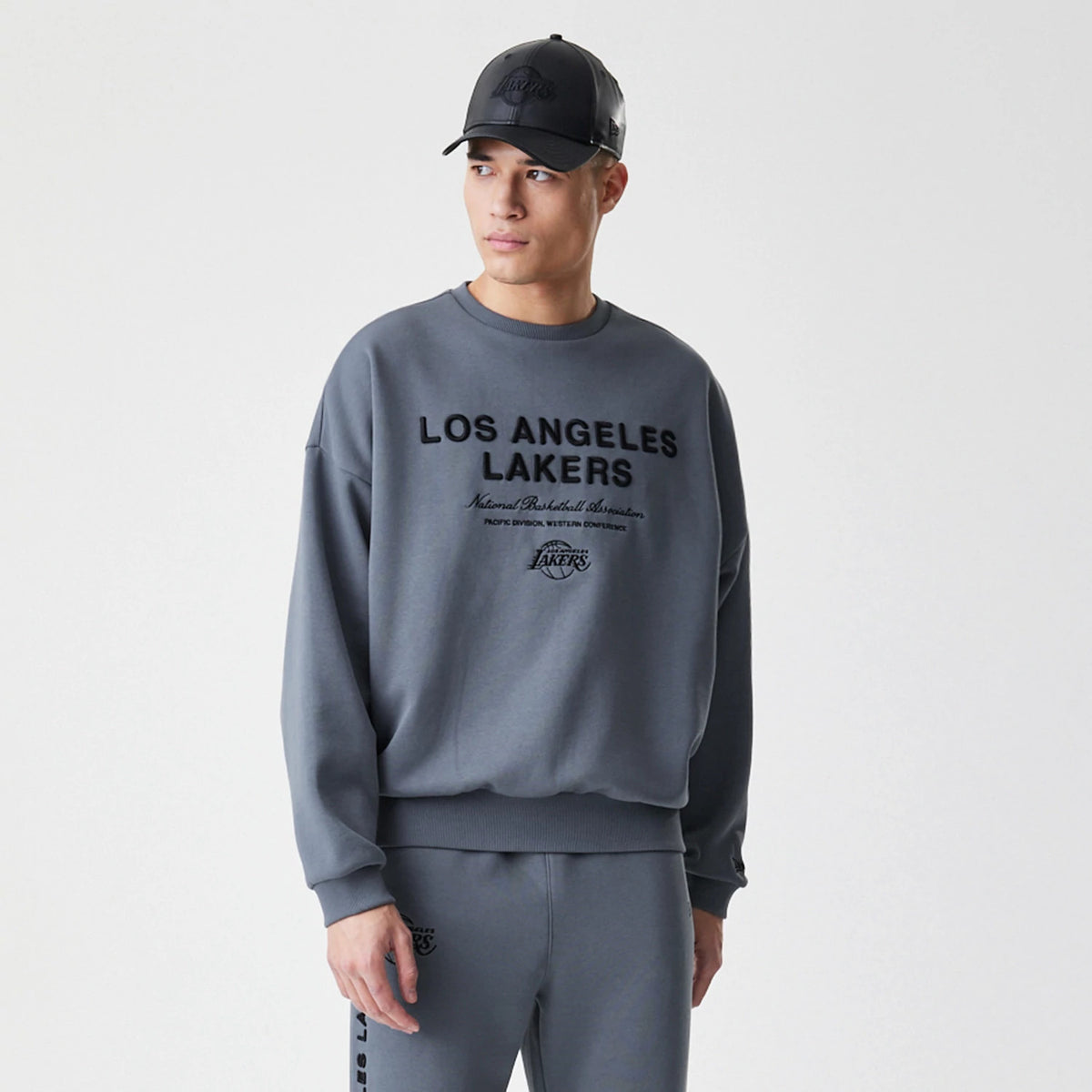 New Era LA Lakers NBA Linear Dark Grey Crew Neck Sweater, front view on a model with a matching dark grey cap, featuring black embroidered Los Angeles Lakers lettering across the chest and ribbed cuffs and hem.