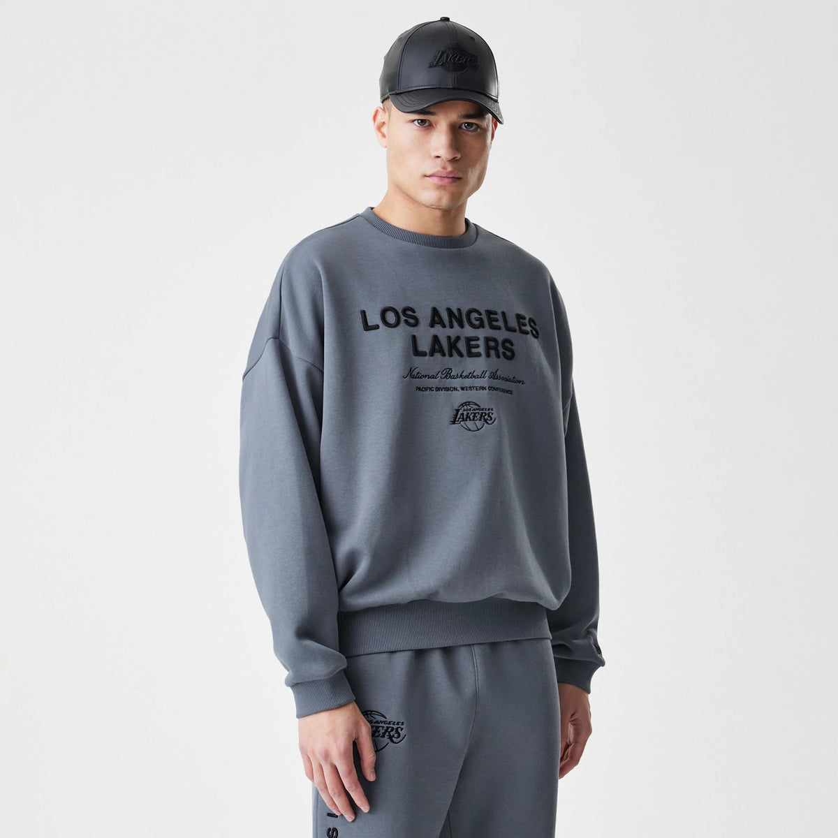 New Era LA Lakers NBA Linear Dark Grey Crew Neck Sweater, front view on a model styled with matching grey joggers, featuring black embroidered Los Angeles Lakers text across the chest and ribbed cuffs and hem.