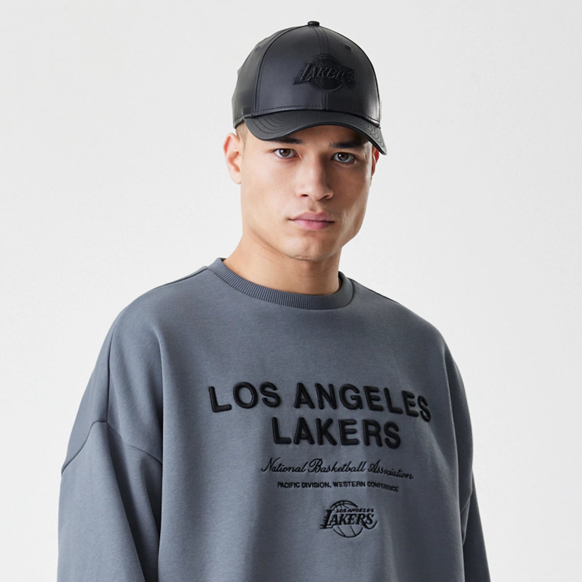 New Era LA Lakers NBA Linear Dark Grey Crew Neck Sweater, front view on a model with matching black cap, featuring Los Angeles Lakers lettering across the chest and additional detailed team text and logo below.
