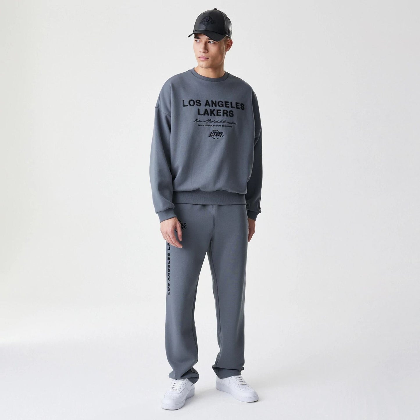 New Era LA Lakers NBA Linear Dark Grey Crew Neck Sweater, front view on a model with matching sweatpants and a black cap, featuring black Los Angeles Lakers lettering across the chest and elasticated cuffs and hem.