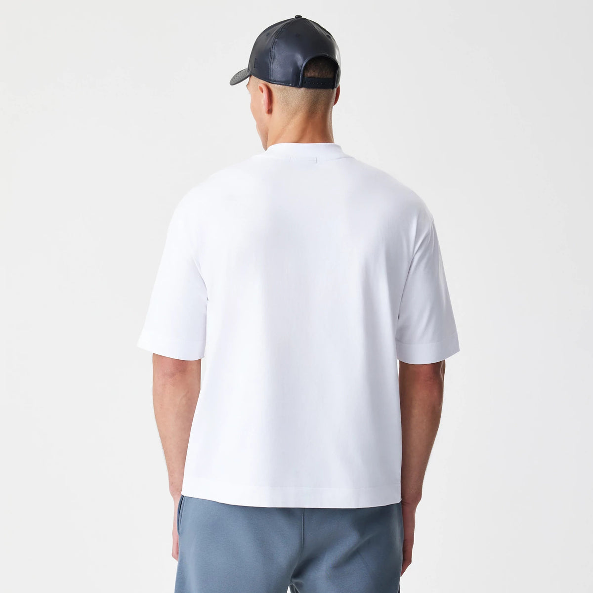 New Era LA Lakers NBA Linear White T-Shirt, back view on-model showing a plain white back with relaxed fit sleeves, styled with a dark cap and grey trousers, highlighting the clean and simple design of the shirt.
