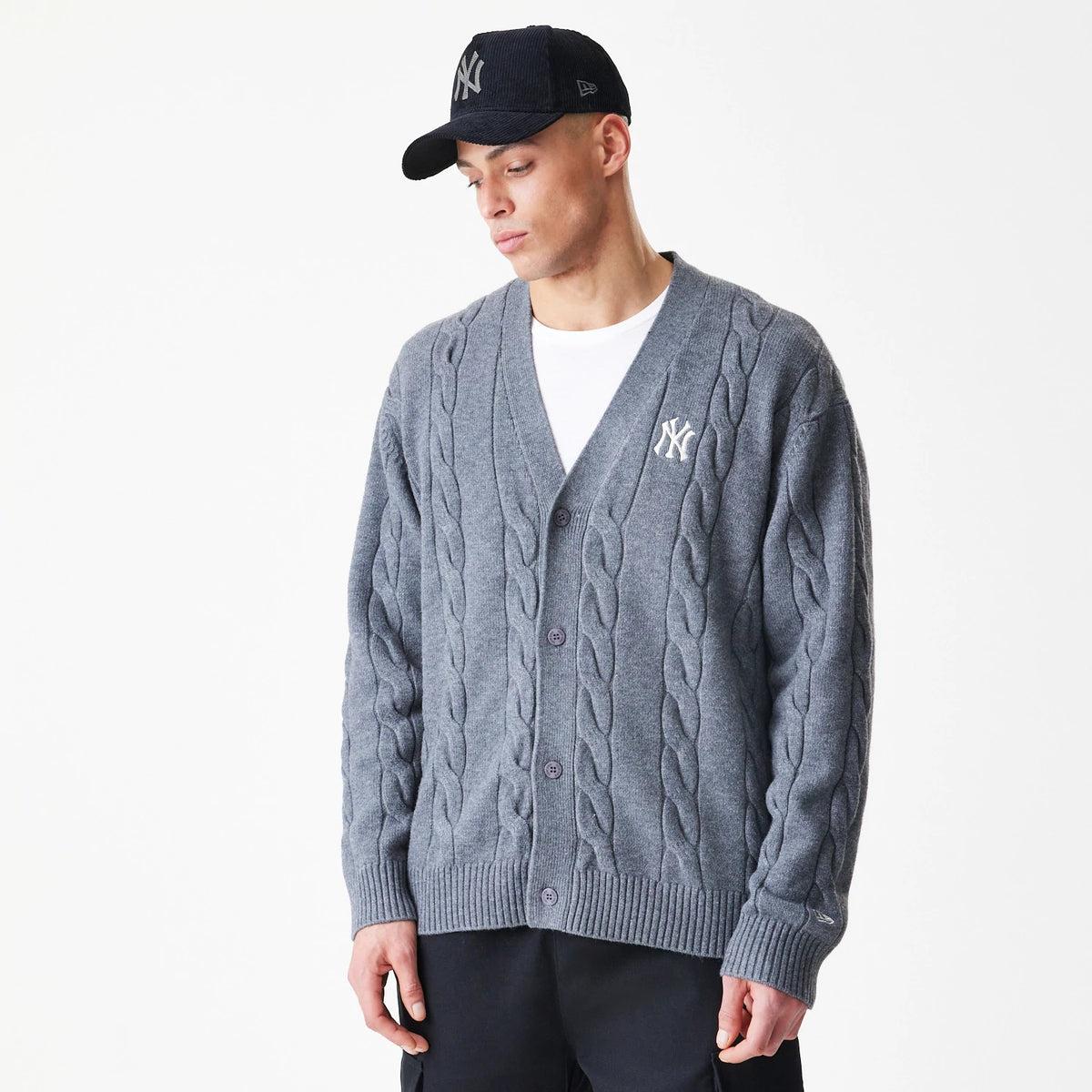 New Era New York Yankees MLB Cable Knit Dark Grey Cardigan, on-model view showing the classic cable knit pattern and embroidered Yankees logo on the left chest, worn over a white T-shirt with dark trousers.