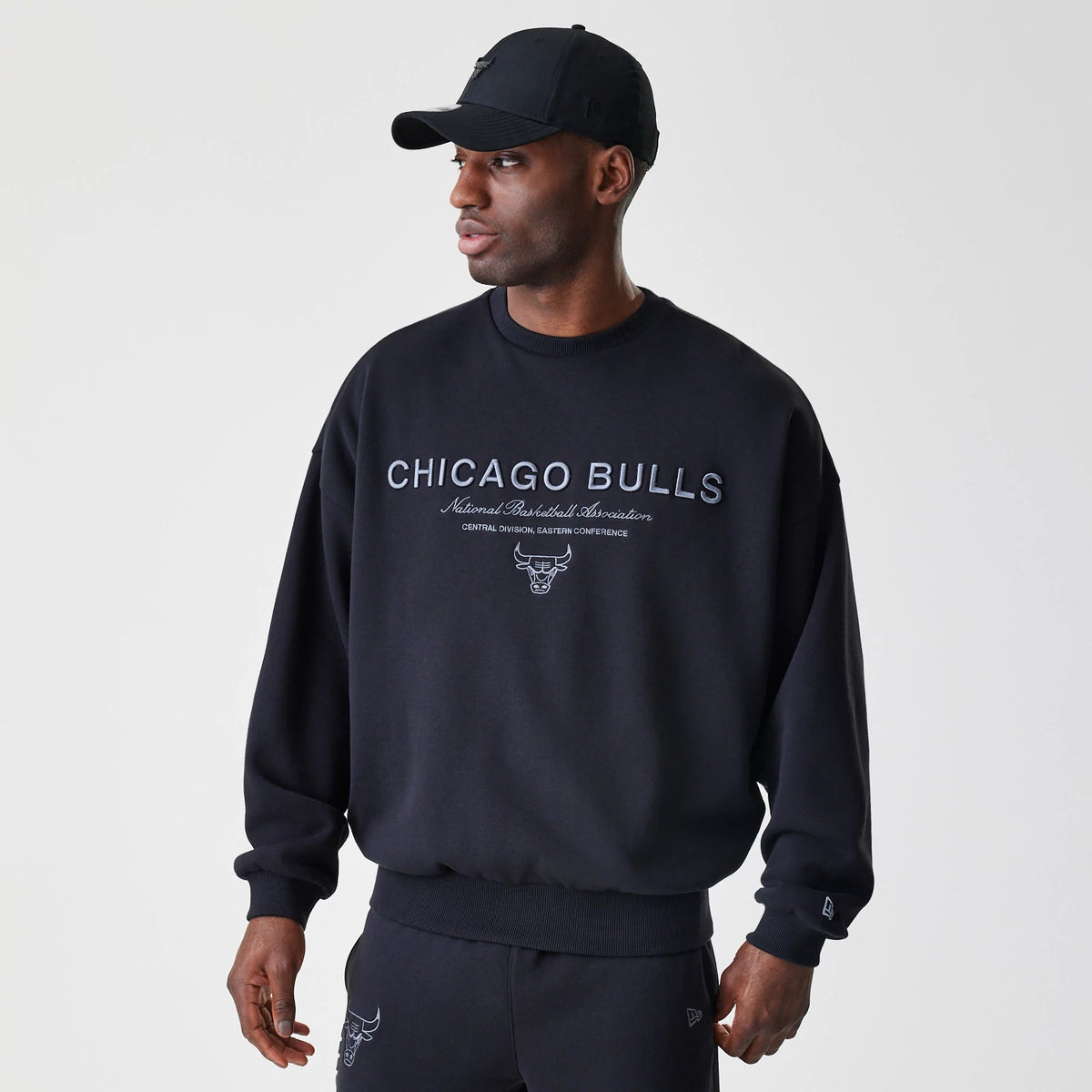 New Era Chicago Bulls NBA Linear Black Crew Neck Sweater, on-model front view with the embroidered Chicago Bulls logo and text across the chest, worn with matching black joggers and a black cap.