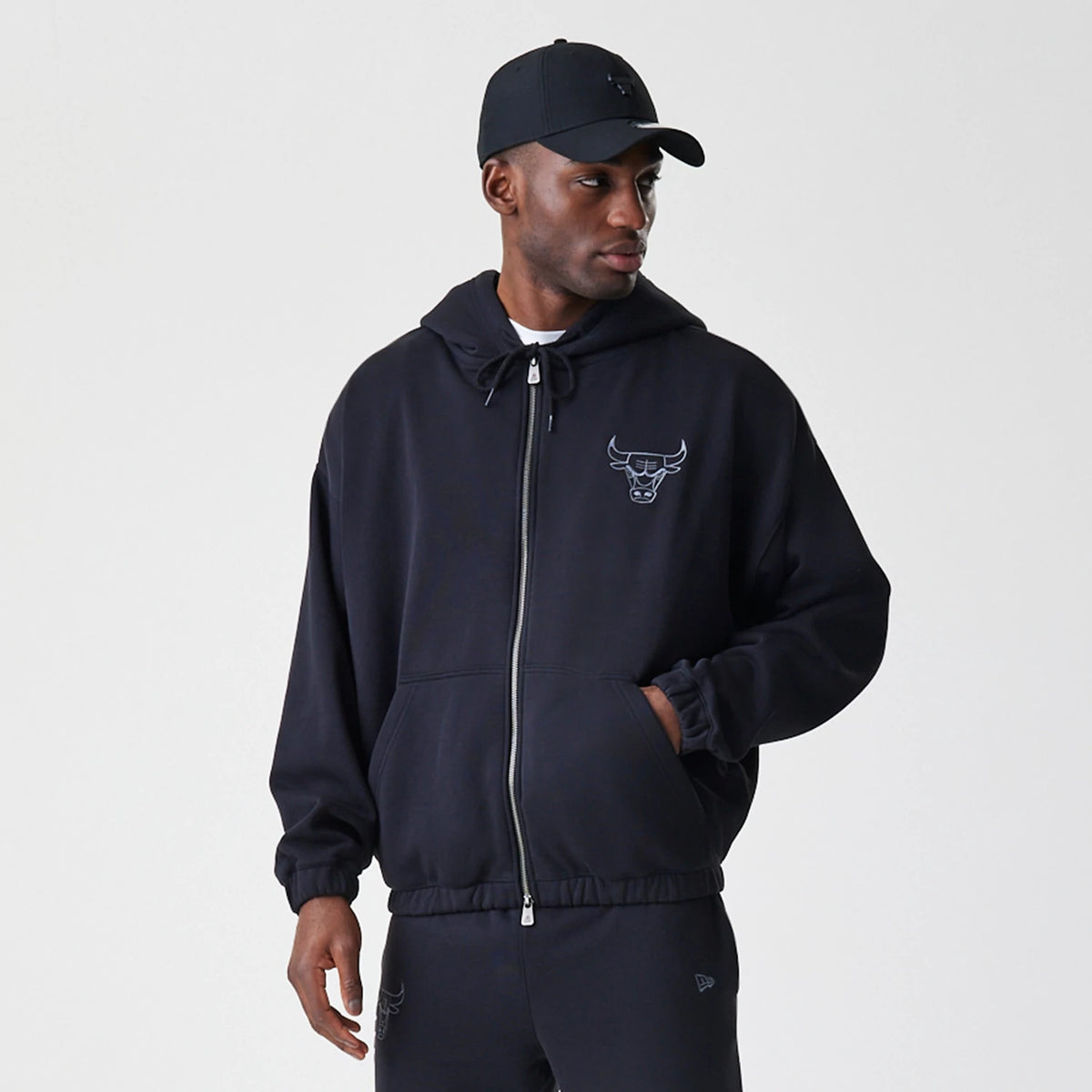New Era Chicago Bulls NBA Linear Black Oversized Full Zip Hoodie, front view on-model showing an embroidered Bulls logo on the left chest, full zip closure and two pockets, styled with matching black trousers and a cap.