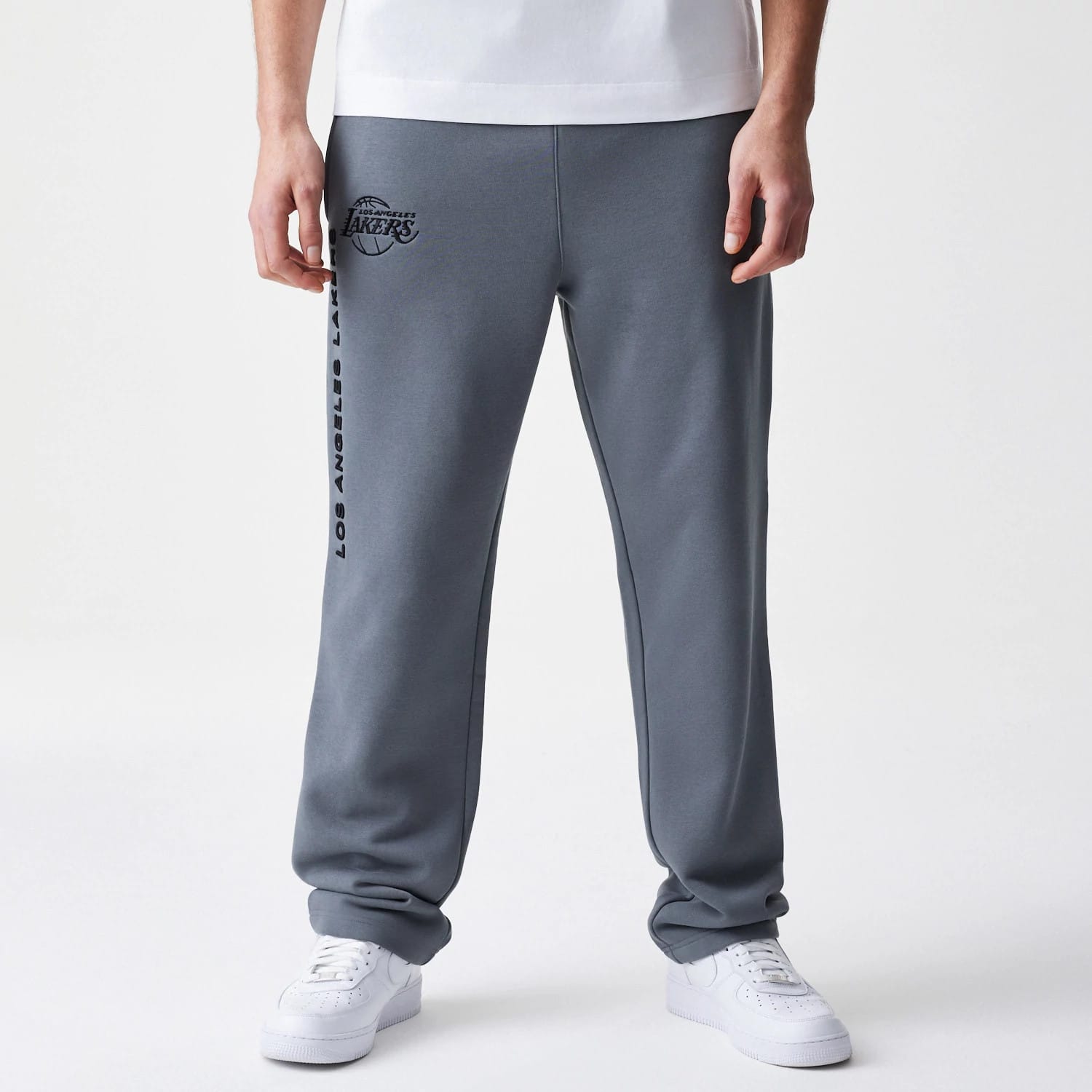 New Era LA Lakers NBA Linear Dark Grey Straight Leg Joggers, front view on a model showing the black LA Lakers logo on the left hip and Los Angeles Lakers lettering running vertically down the right leg, paired with white trainers.