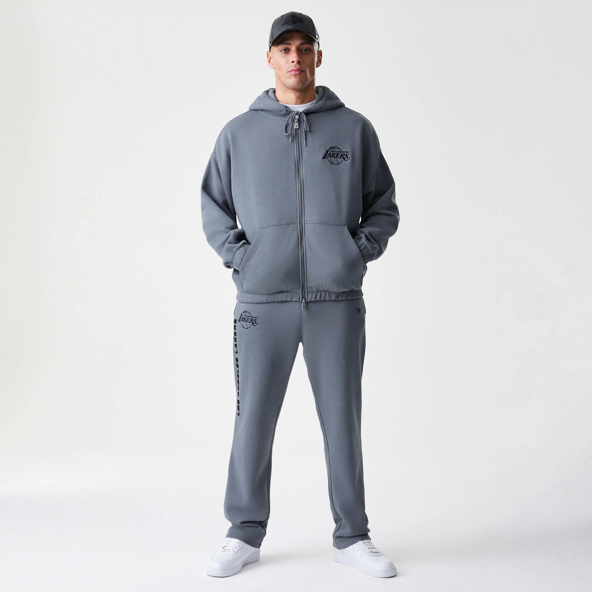 New Era LA Lakers NBA Linear Dark Grey Straight Leg Joggers, front view on a model with matching hoodie and cap, featuring the Lakers logo on the left thigh and linear lettering running down the right leg.