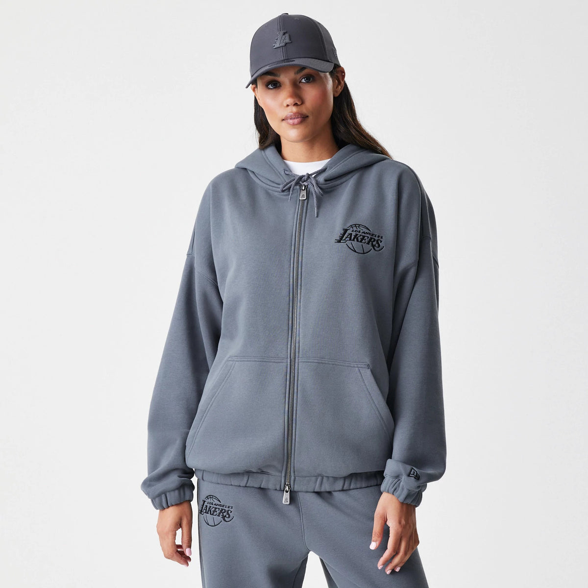 New Era LA Lakers NBA Linear Dark Grey Oversized Full Zip Hoodie, front view on a model with a matching cap and joggers, showing an embroidered LA Lakers logo on the left chest and a full zip closure down the centre.