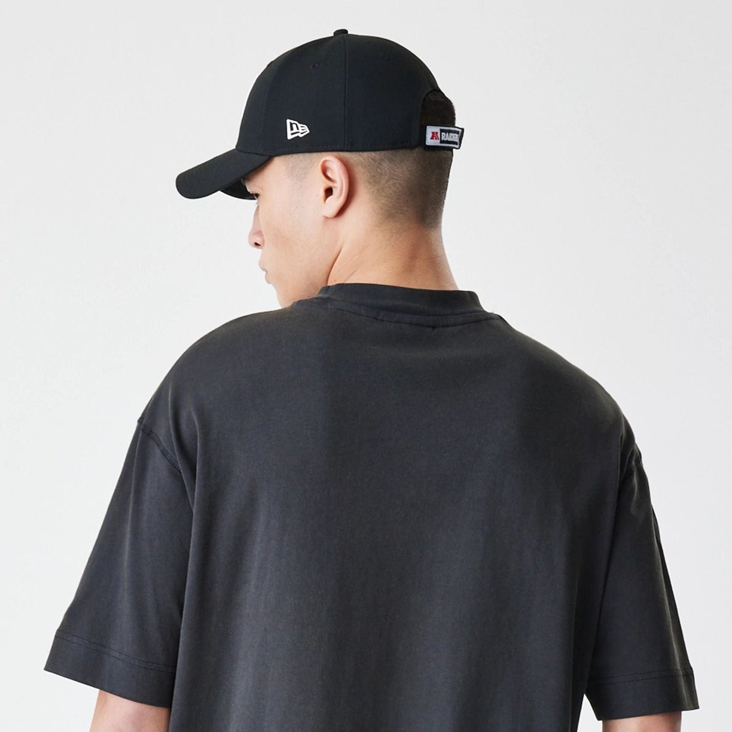 New Era Las Vegas Raiders NFL Washed Boxy Dark Grey Oversized T-Shirt, back view on a model also wearing a cap, showing the dark grey fabric extending loosely across the shoulders with short sleeves and no logos or graphics.