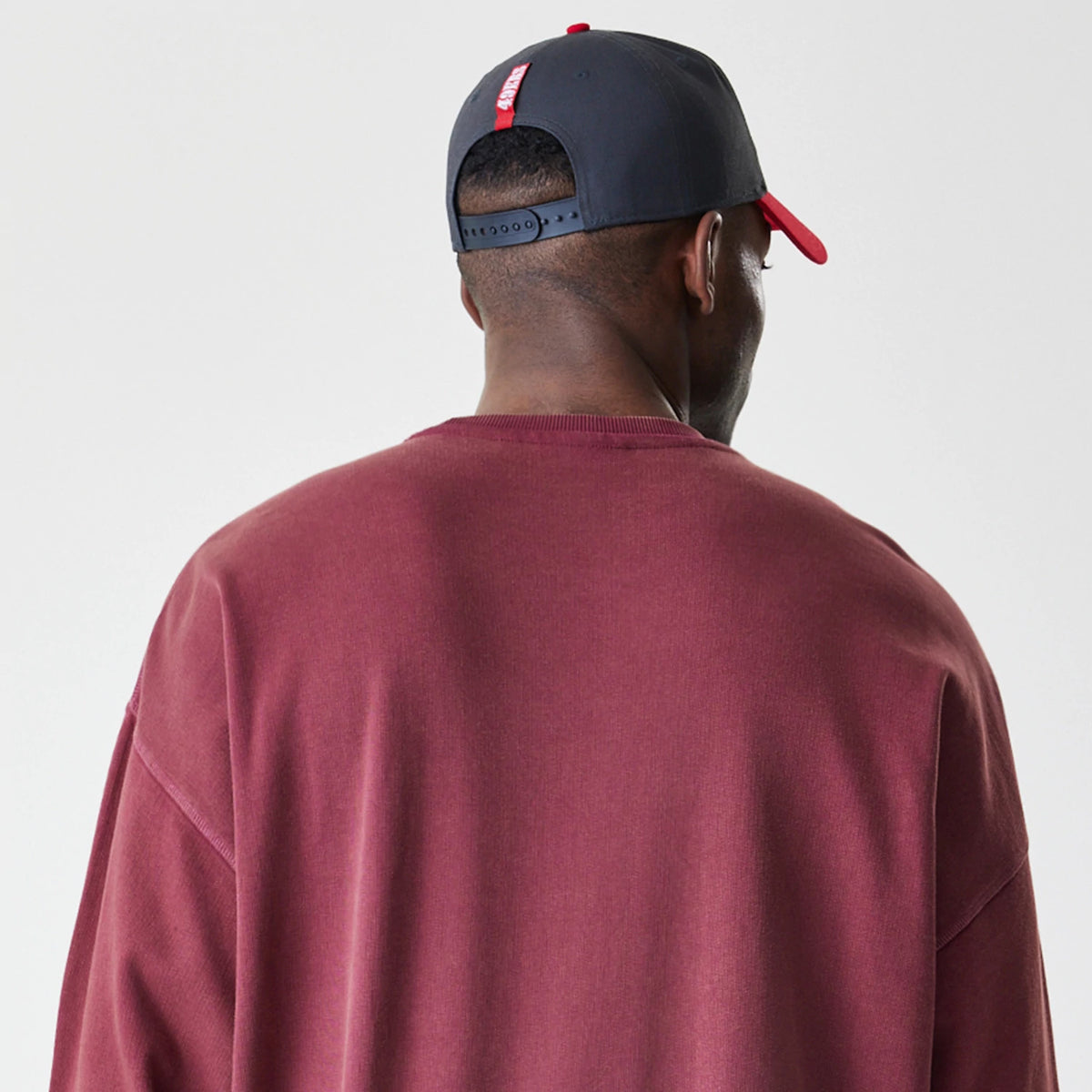New Era San Francisco 49ers NFL Washed Boxy Dark Red Oversized Crew Neck Sweater, back view on a model with a black snapback cap, showing the relaxed fit and ribbed neckline of the sweater in dark red fabric.