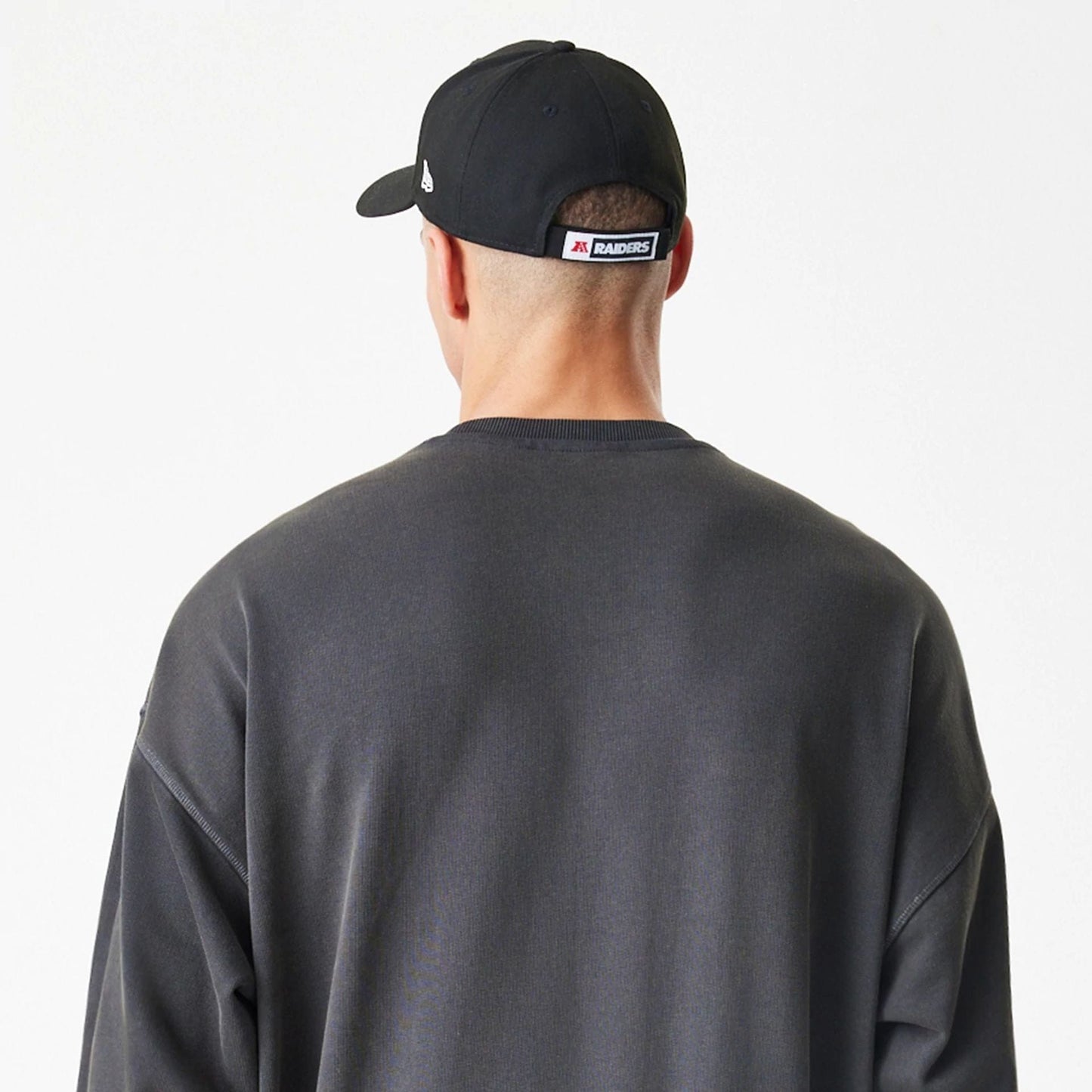 New Era Las Vegas Raiders NFL Washed Boxy Dark Grey Oversized Crew Neck Sweater, back view on a model wearing a black cap, displaying the plain dark grey fabric and relaxed fit with dropped shoulders.