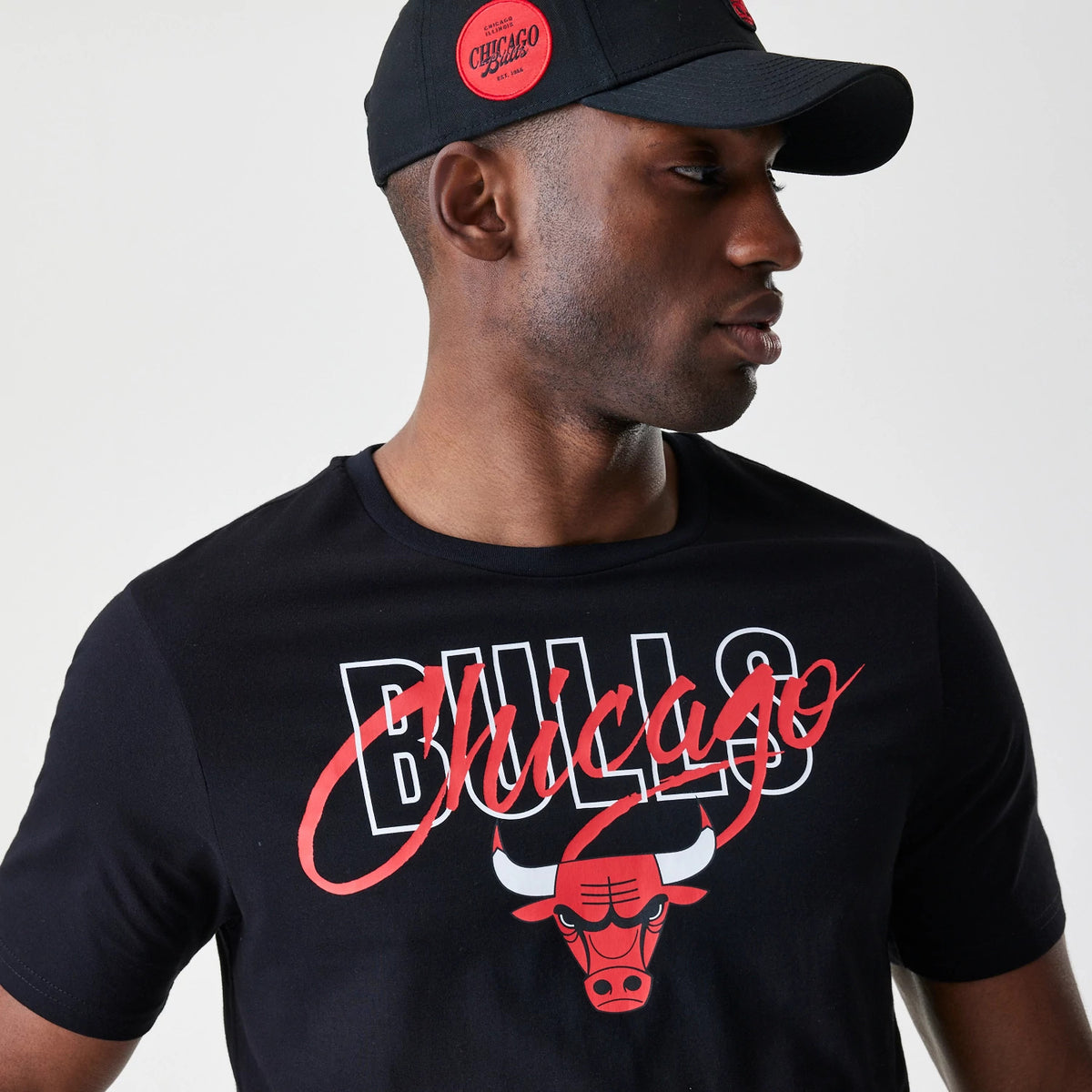 New Era Chicago Bulls NBA Script Black T-Shirt, front view showing a graphic with red Chicago script over large white Bulls lettering and the team's red bull logo, worn with a coordinating black cap featuring a red round patch.