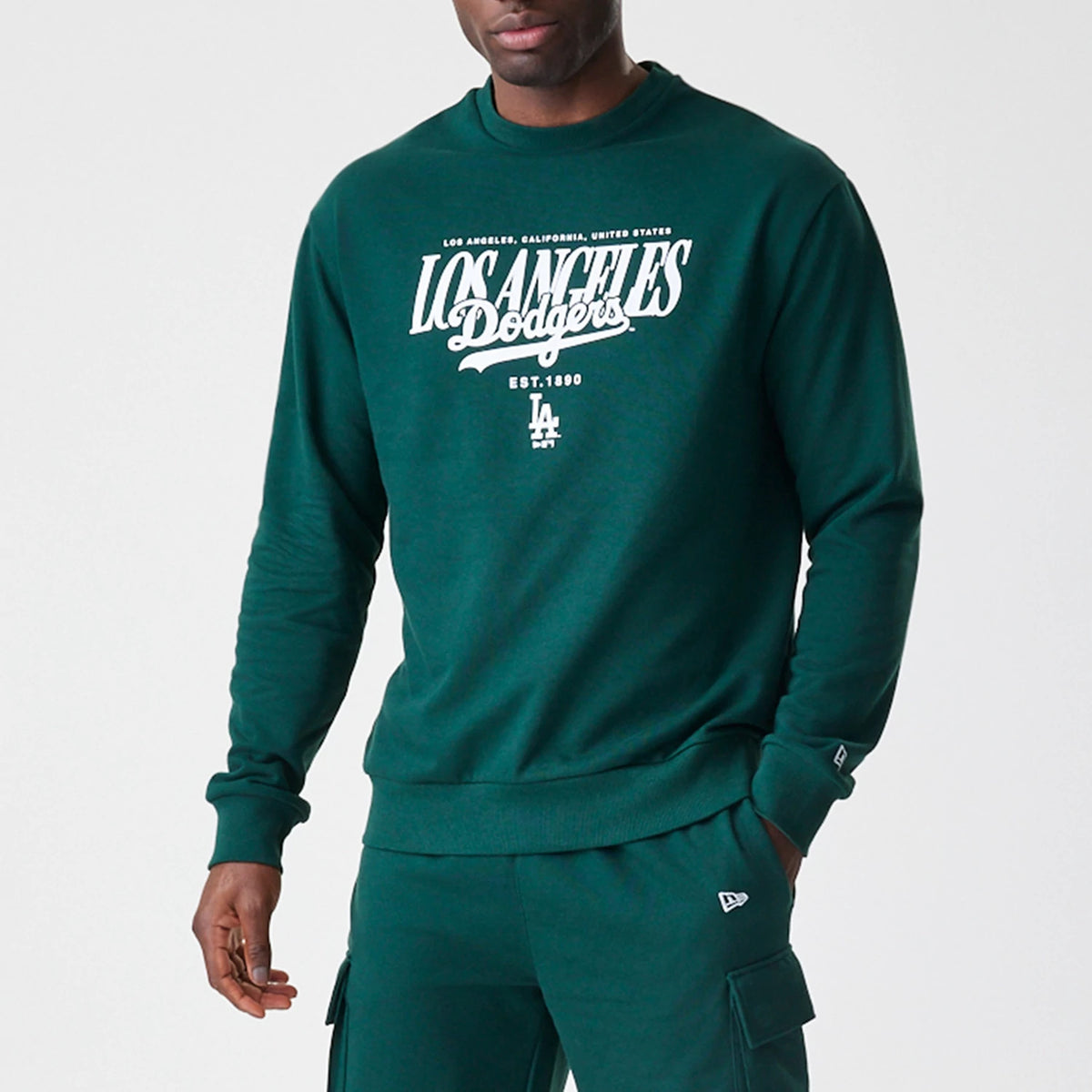 New Era LA Dodgers MLB Script Dark Green Crew Neck Sweater, front view on a model with white printed script and Dodgers logo across the chest, featuring a small New Era flag logo on the left sleeve.