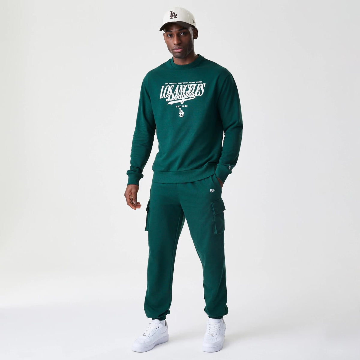New Era LA Dodgers MLB Script Dark Green Crew Neck Sweater, front view on a model styled with matching trousers, showing the white embroidered Los Angeles Dodgers script logo across the chest and New Era branding on the left sleeve.