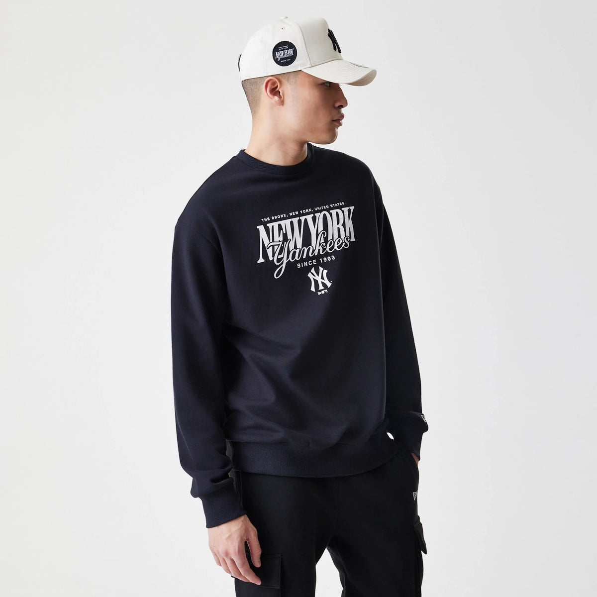 New Era New York Yankees MLB Script Black Crew Neck Sweater, on-model showing a white New York Yankees logo with script lettering on the front, styled with a light-coloured cap featuring an NY logo.