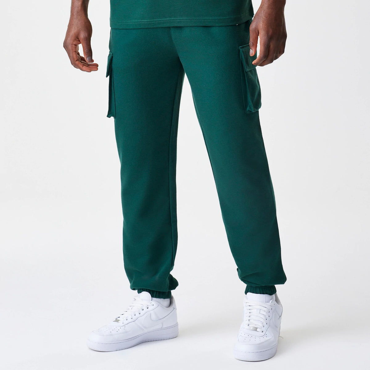 New Era Core Cargo Dark Green Joggers, front view on a model showing the relaxed fit with practical side cargo pockets and elasticated cuffs, paired with white trainers for a casual look.