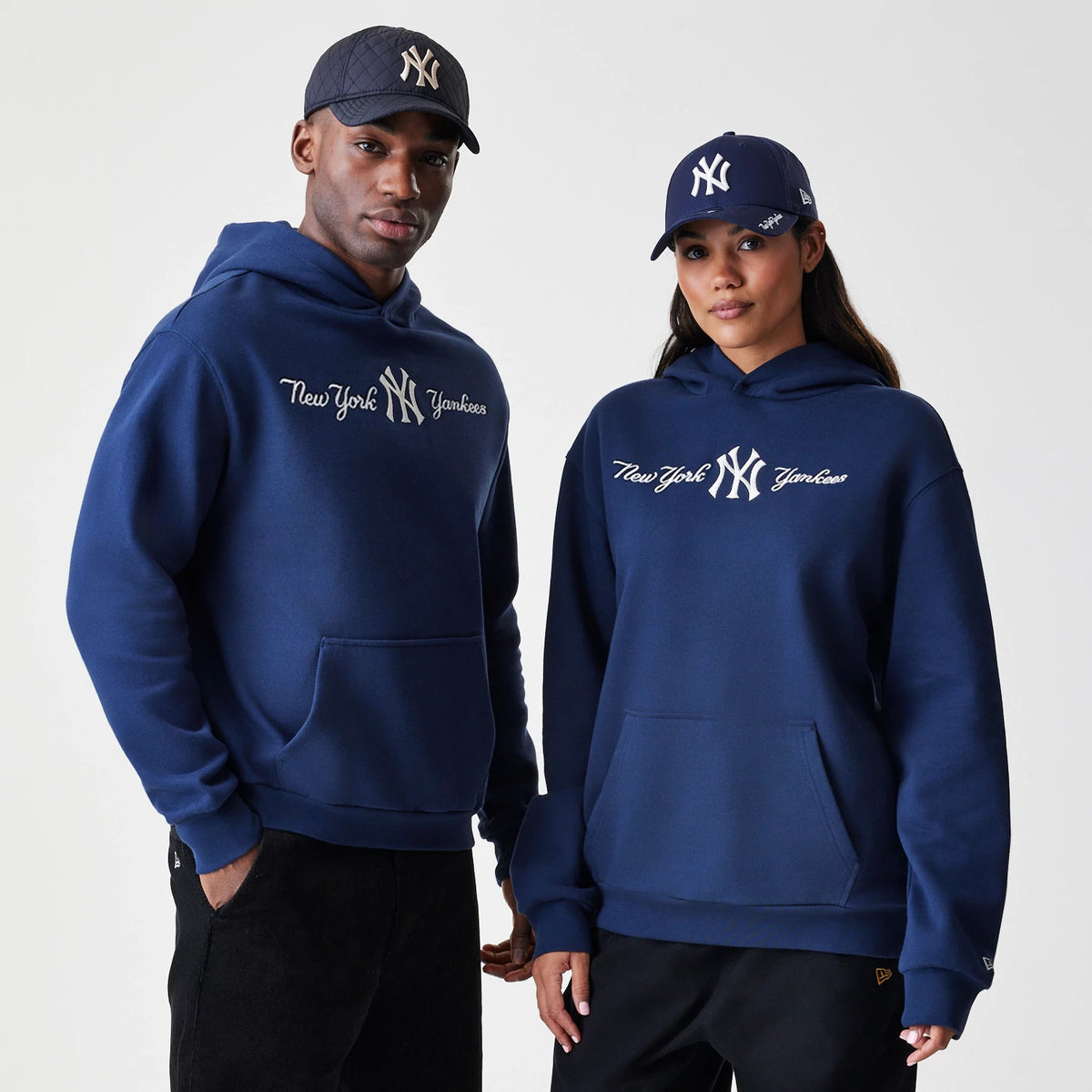 New Era New York Yankees MLB Script Navy Oversized Pullover Hoodie, front view on two models showing the embroidered New York Yankees script and logo across the chest, paired with matching navy caps displaying an embroidered NY logo.