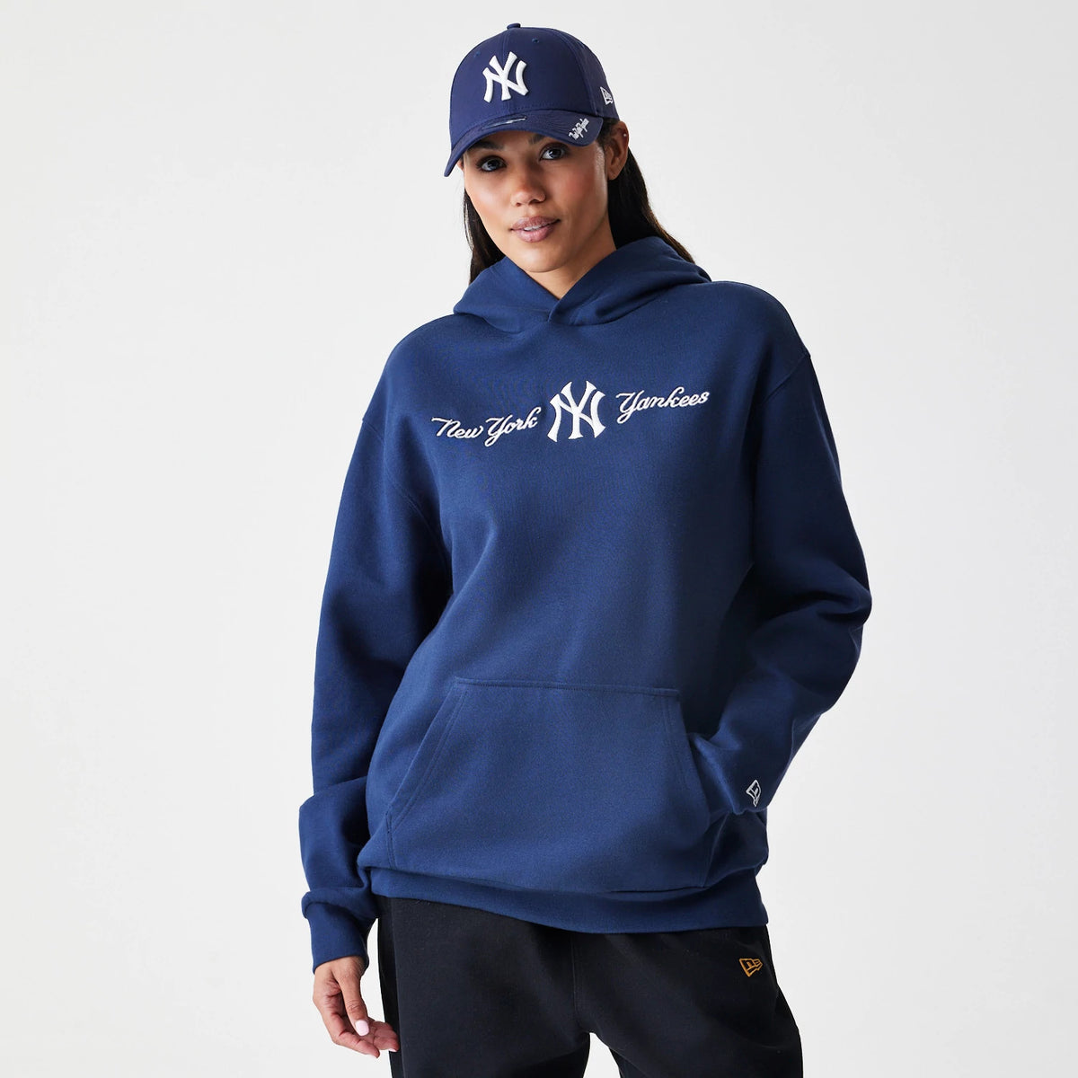 New Era New York Yankees MLB Script Navy Oversized Pullover Hoodie, front view on a model with a matching cap, showing the white embroidered New York Yankees logo and script across the chest centre, an open kangaroo pocket below.
