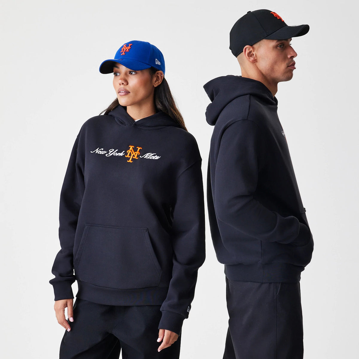 New Era New York Mets MLB Script Black Oversized Pullover Hoodie, front view on models, featuring New York Mets embroidered script and logo in white and orange on the chest, styled with a blue cap and a black cap.