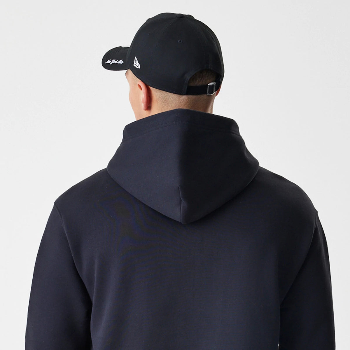 New Era New York Mets MLB Script Black Oversized Pullover Hoodie, back view on a model wearing a matching cap, showing plain black fabric with a hood folded down over the upper back.